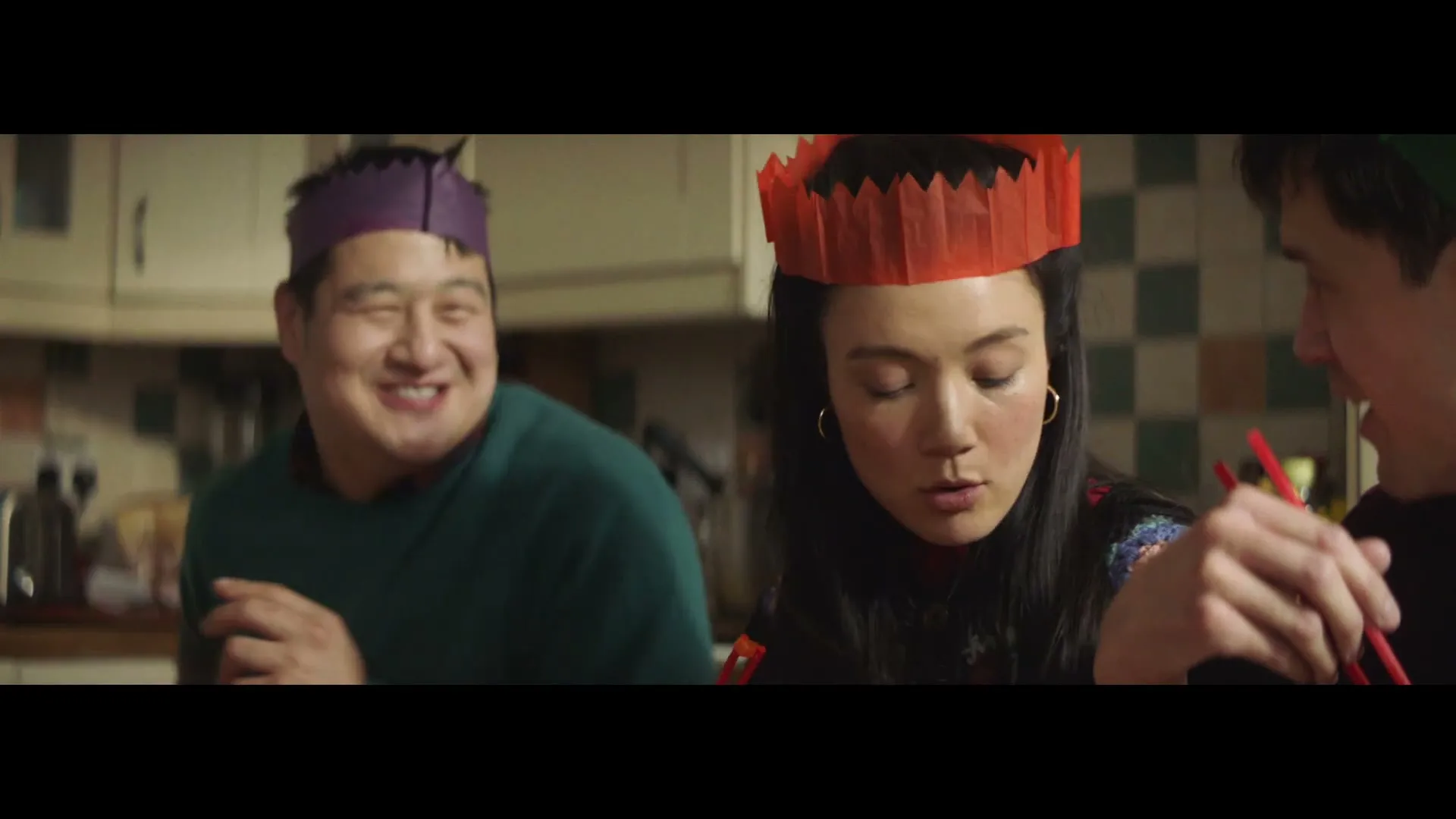 A woman in a red paper crown looks down while two men, one in a purple paper crown, eat with chopsticks at a kitchen table.