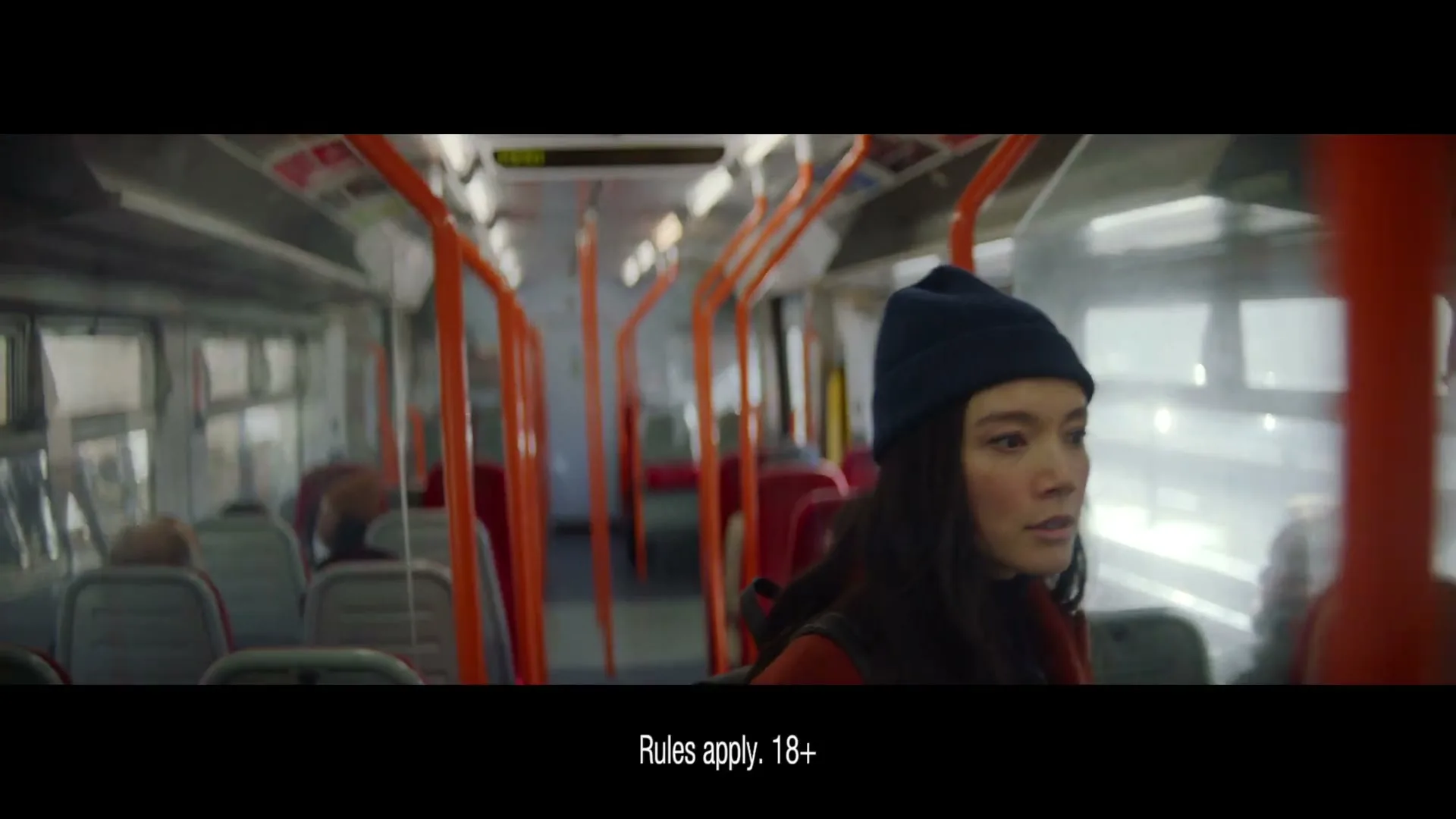 A young woman with a blue beanie and red jacket looks out the window of a moving train, with other passengers visible in the background.