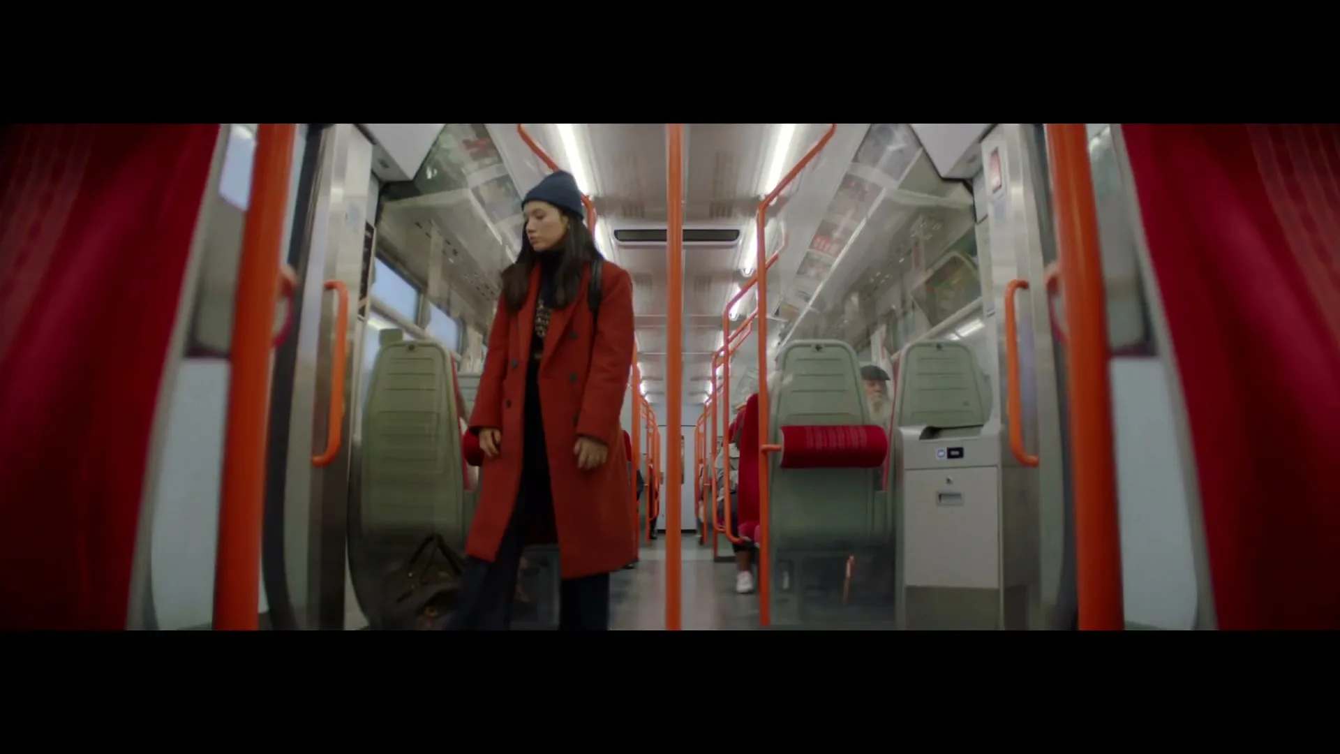 A young woman in a red coat and blue beanie enters a tram, places her bag down, and sits on a seat, looking around.