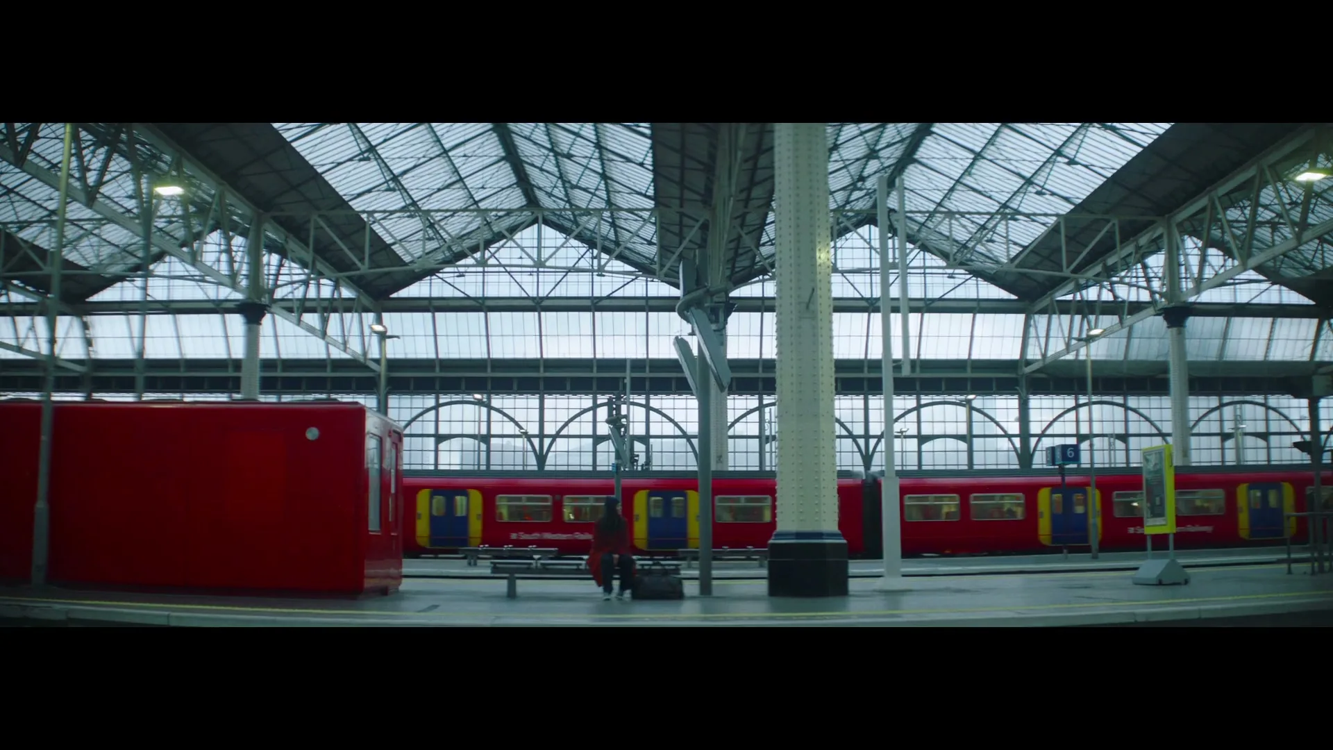 A wide shot shows a woman in a red coat standing on a train platform as a red train pulls away from the station.