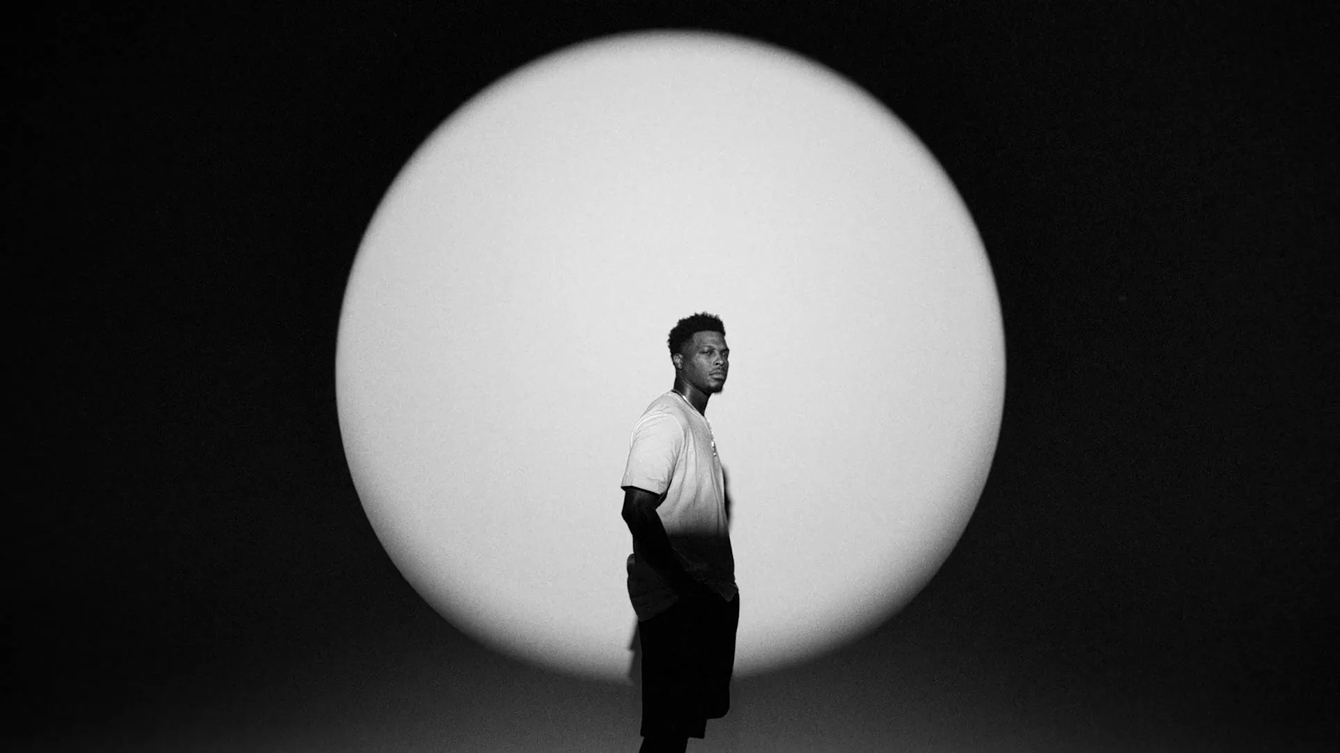 A young Black man in a t-shirt and shorts stands in a spotlight against a dark background, looking to the side.