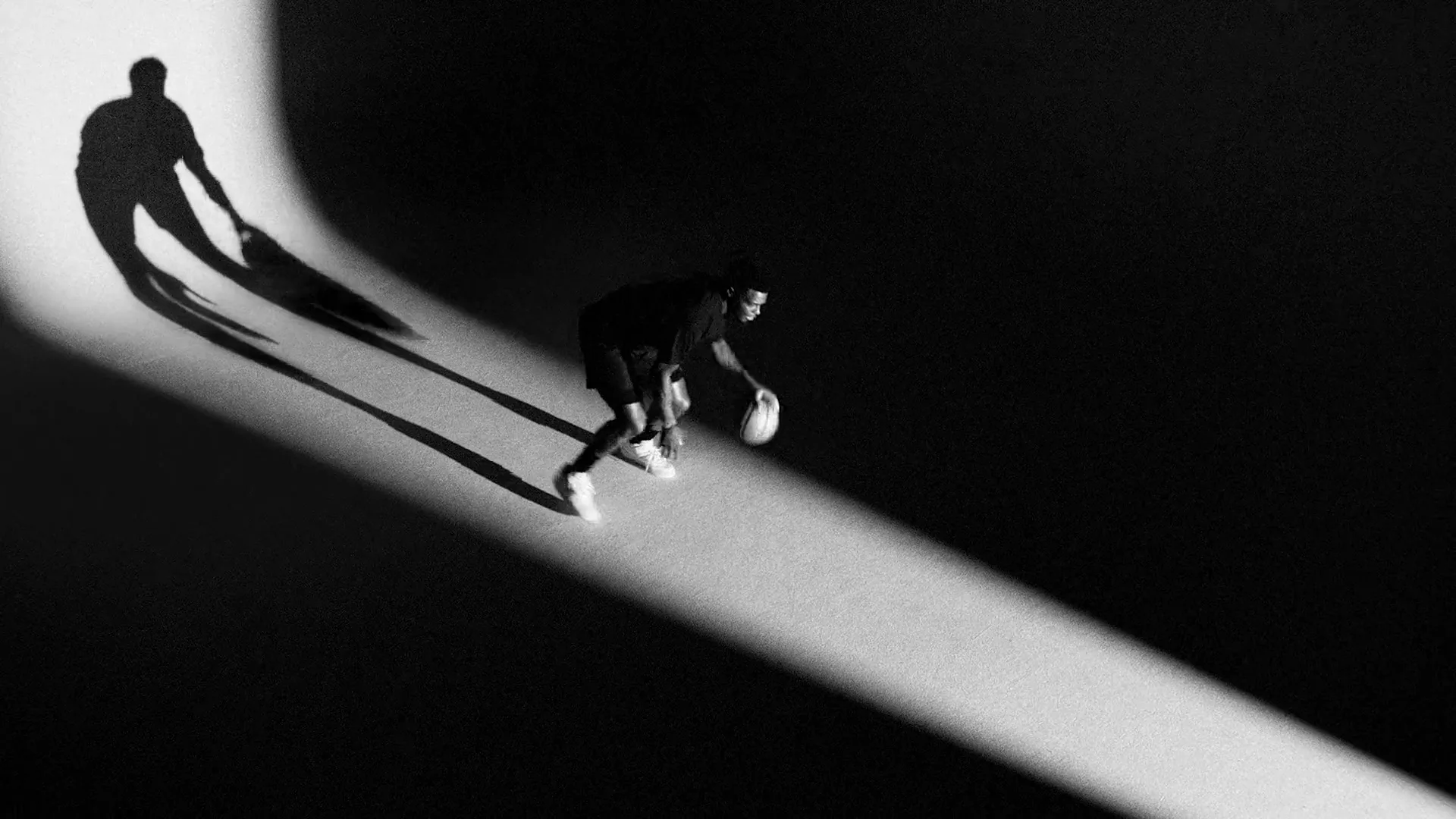 A male basketball player in dark clothing dribbles a basketball in a spotlight, casting a long shadow on a dark floor.