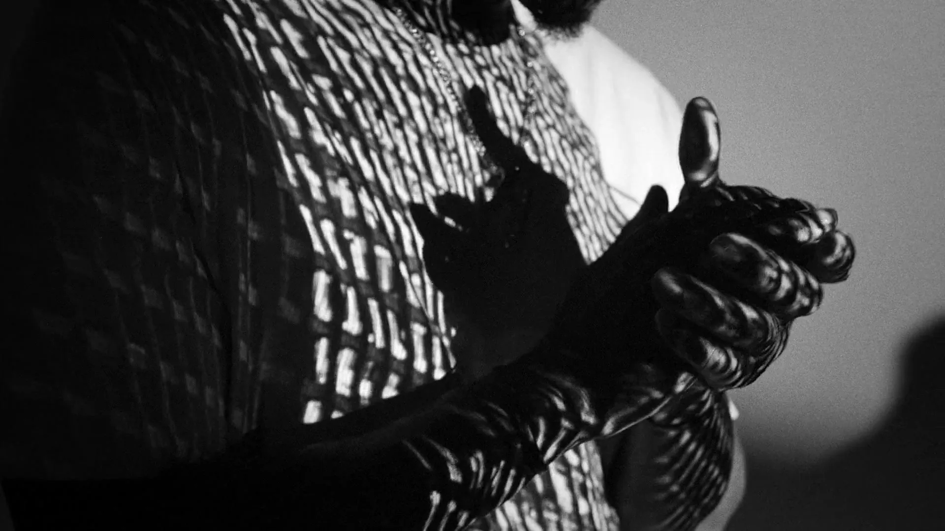 A person in a patterned shirt with a chain necklace claps their hands, with light and shadow creating a striped pattern on their body.