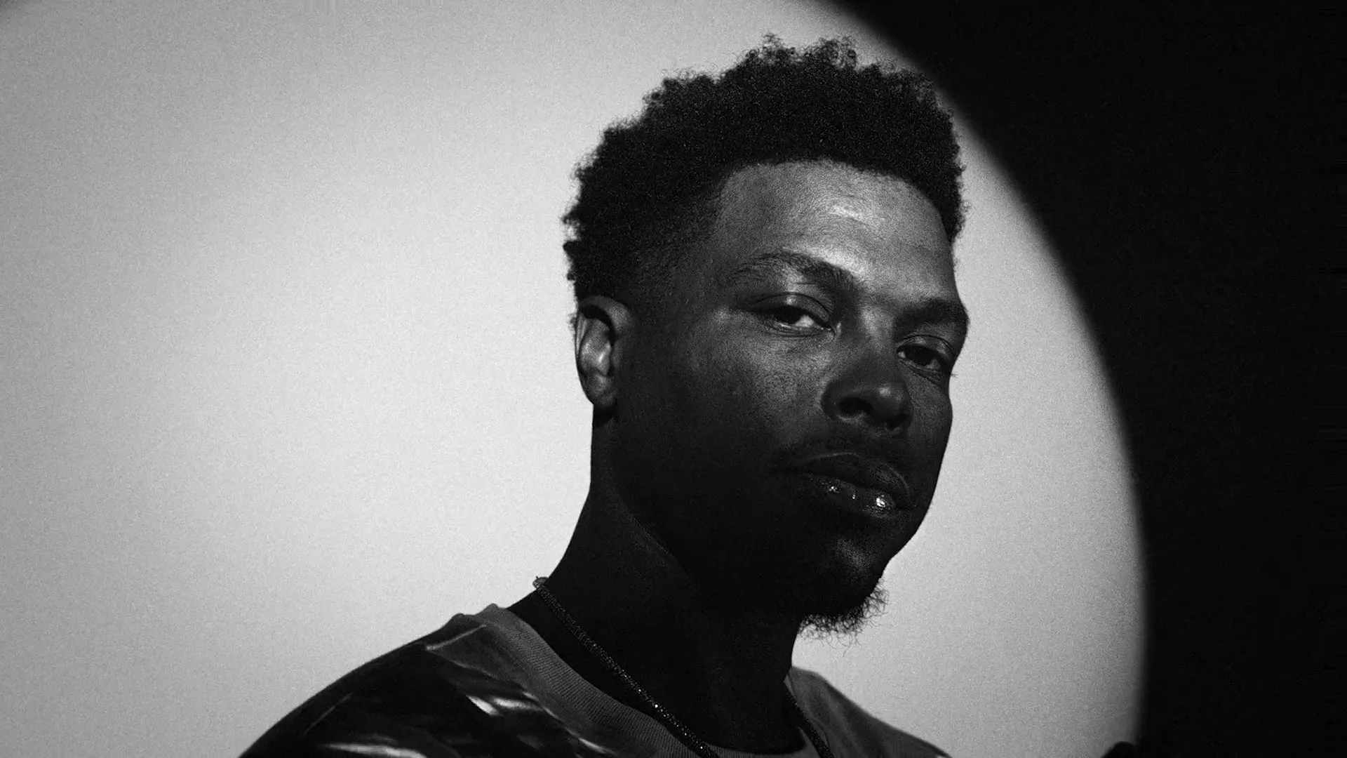 A Black man with a beard and short curly hair looks directly at the camera in a black and white medium closeup shot.