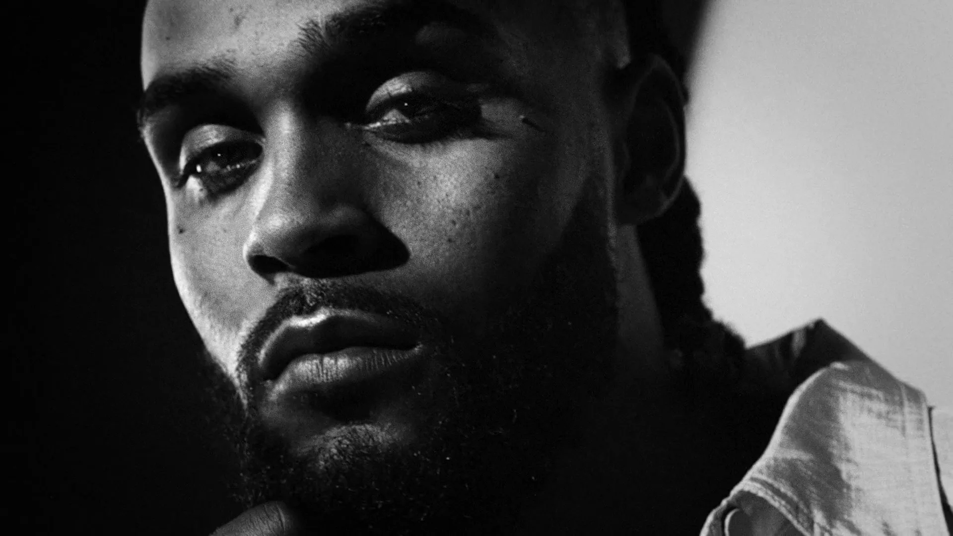 A Black man with a beard and braided hair looks down thoughtfully in a black and white, low-key shot.