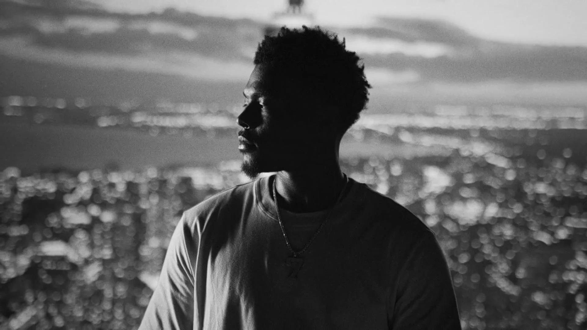 A young Black man with an afro and beard, wearing a t-shirt and necklace, looks out over a blurry city skyline at night, in a black and white shot.