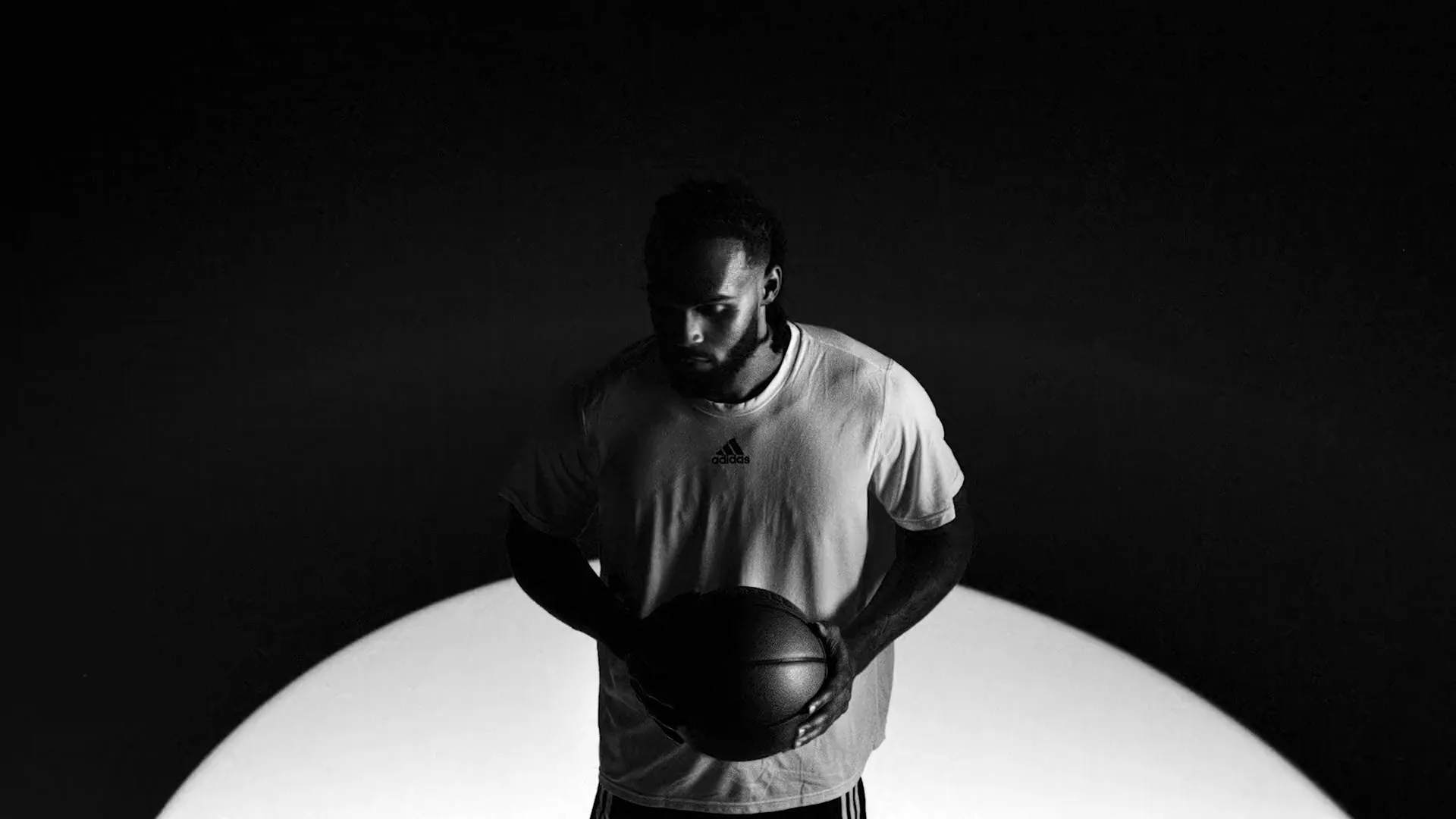 A male basketball player in a t-shirt holds a basketball, looking down in a studio with a spotlight on the floor.