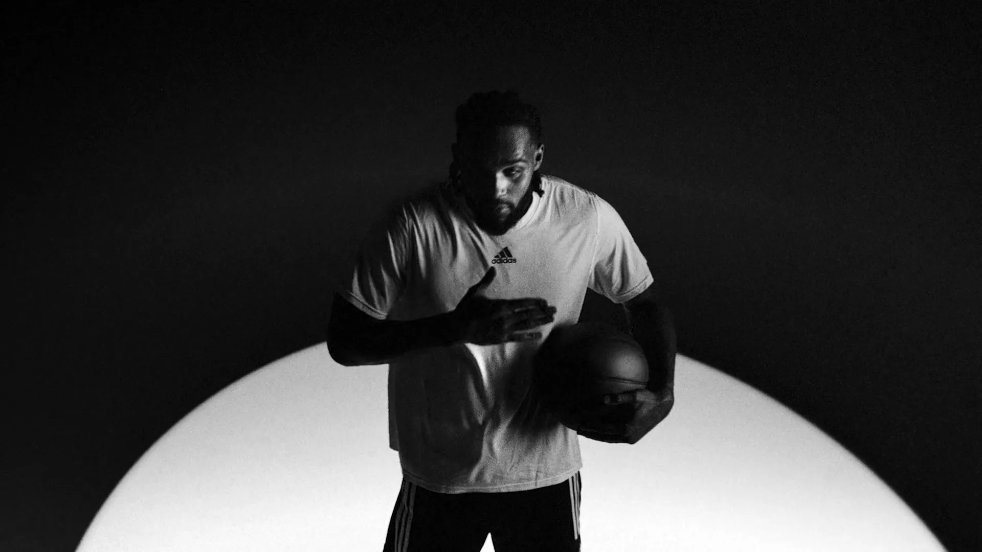 A male basketball player in a white t-shirt holds a basketball and looks up, standing in a spotlight against a dark background.