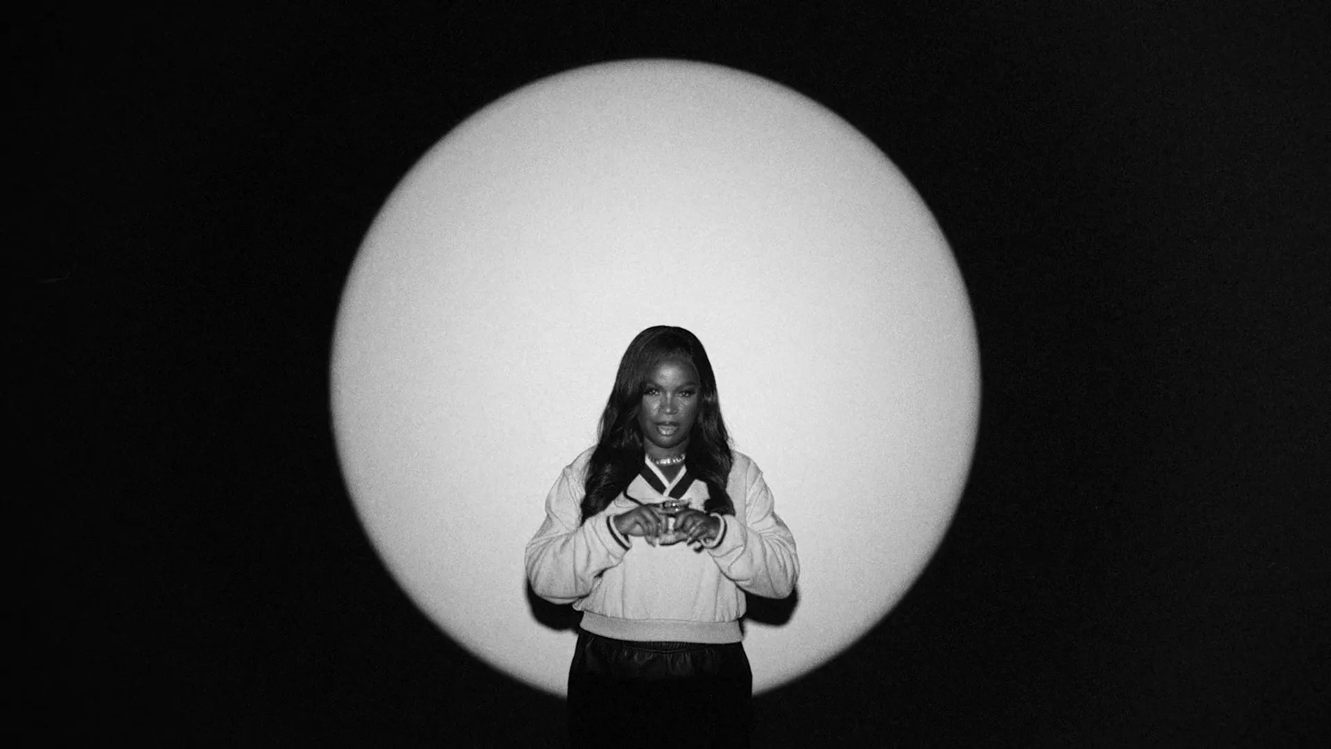 A young Black woman with long dark hair stands in a spotlight, wearing a light-colored sweatshirt and dark pants, moving her hands in a studio void.