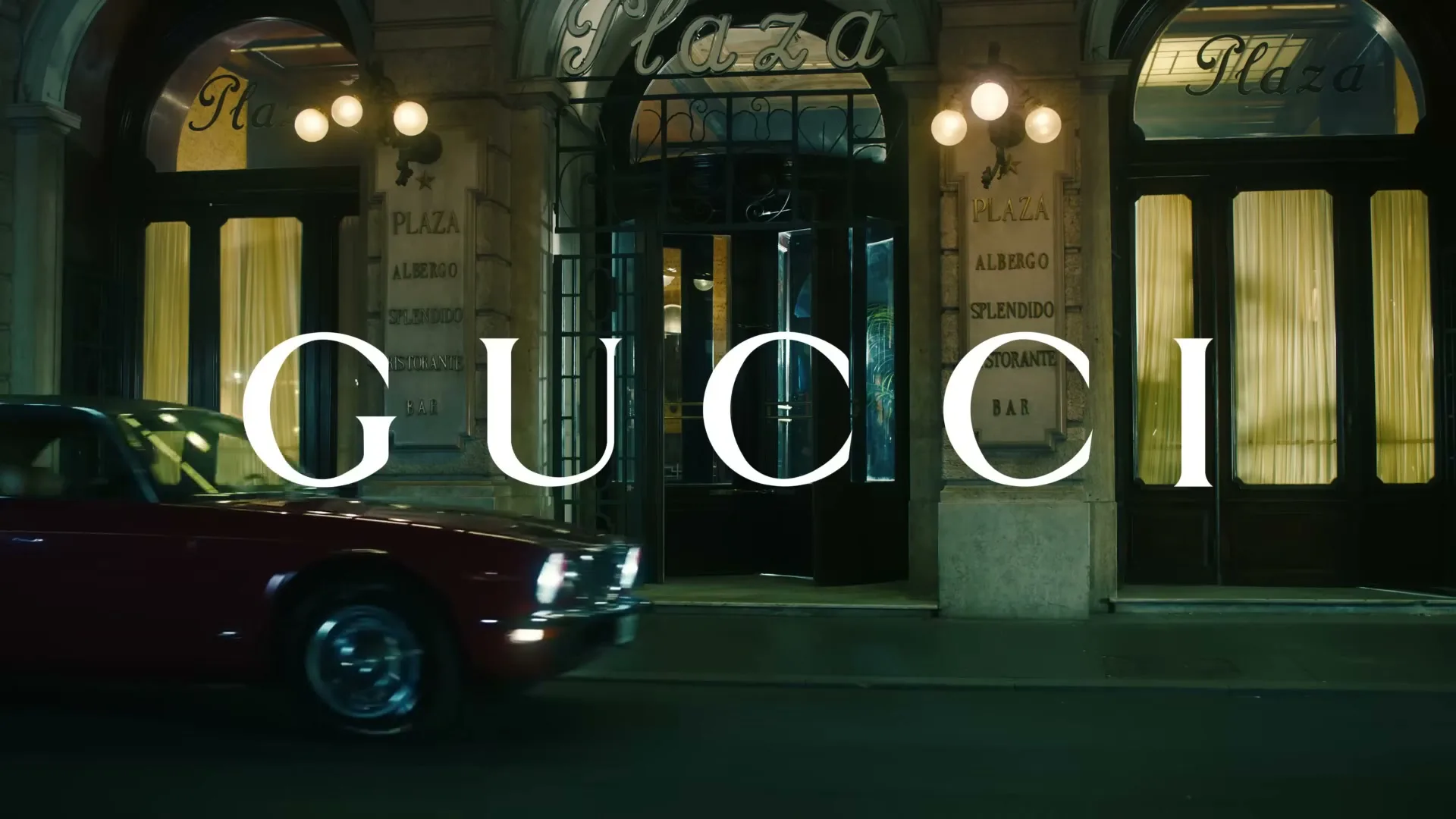 A red vintage car drives past a grand hotel entrance at night, with the word "GUCCI" superimposed in white letters.