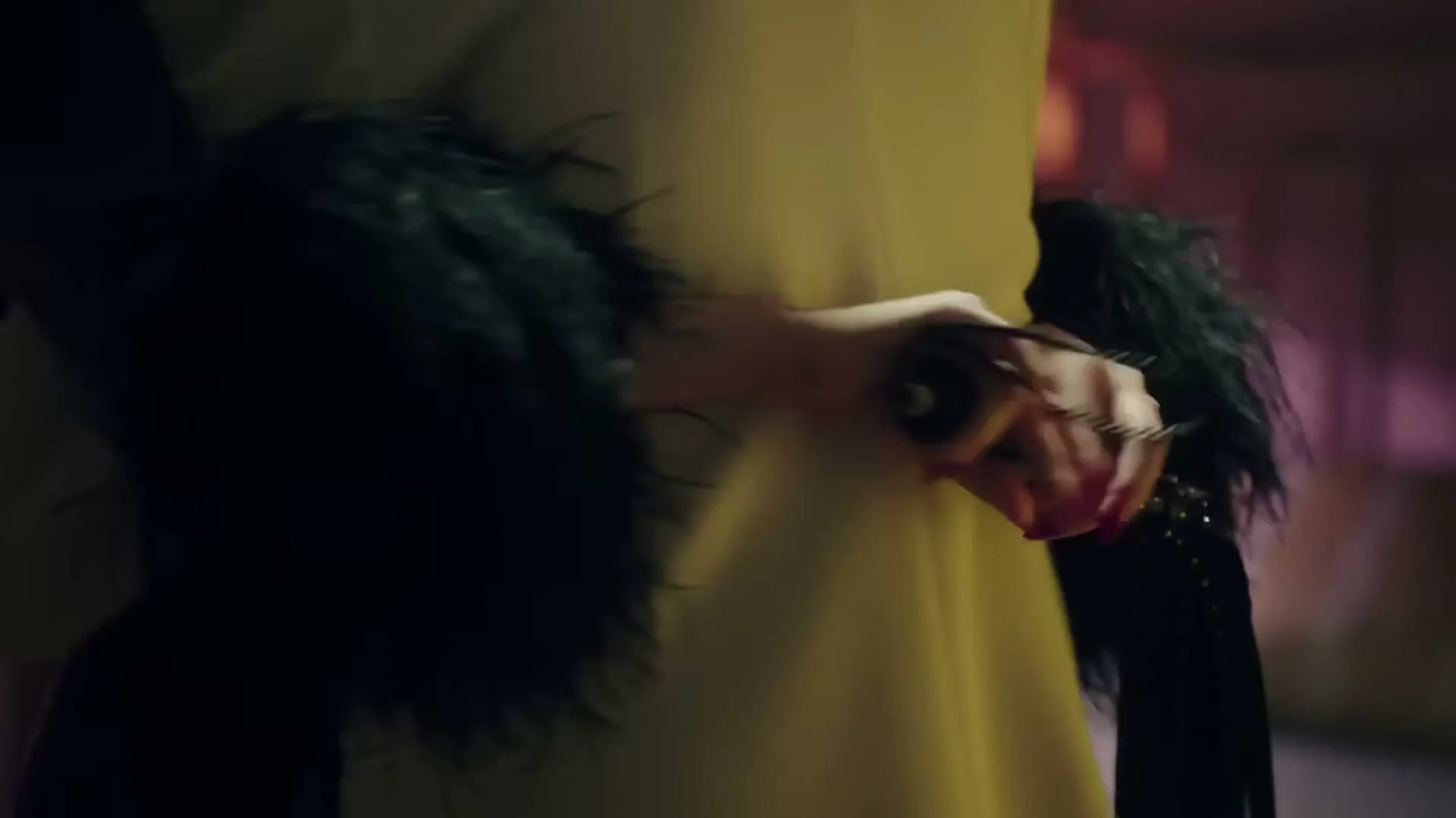 A person in a yellow dress with black feathered sleeves holds a black studded leash in their hand, in an interior setting.