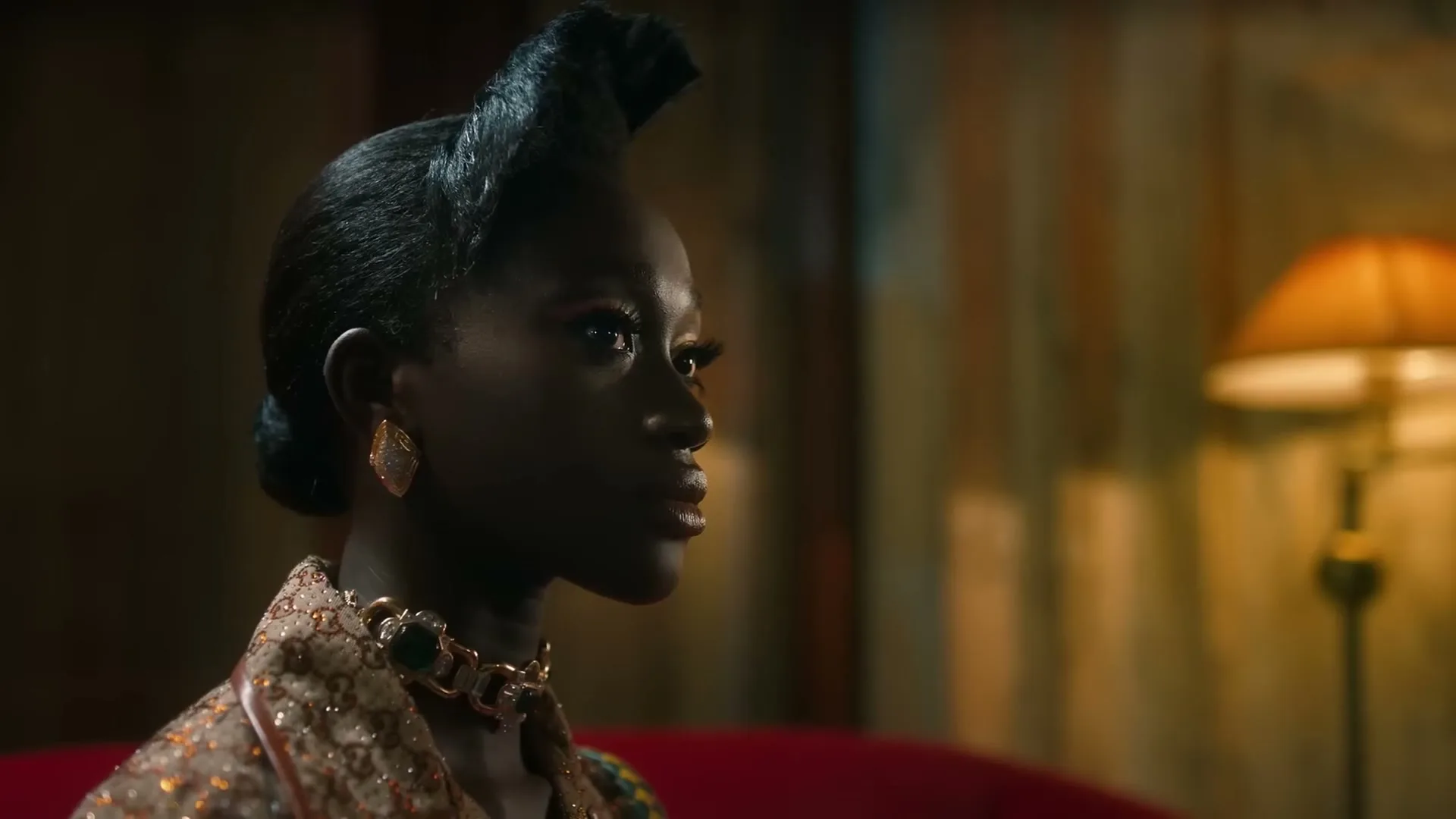 A Black female model with an elaborate hairstyle and ornate jewelry looks off-camera, then turns to face the viewer in a dimly lit, opulent room.