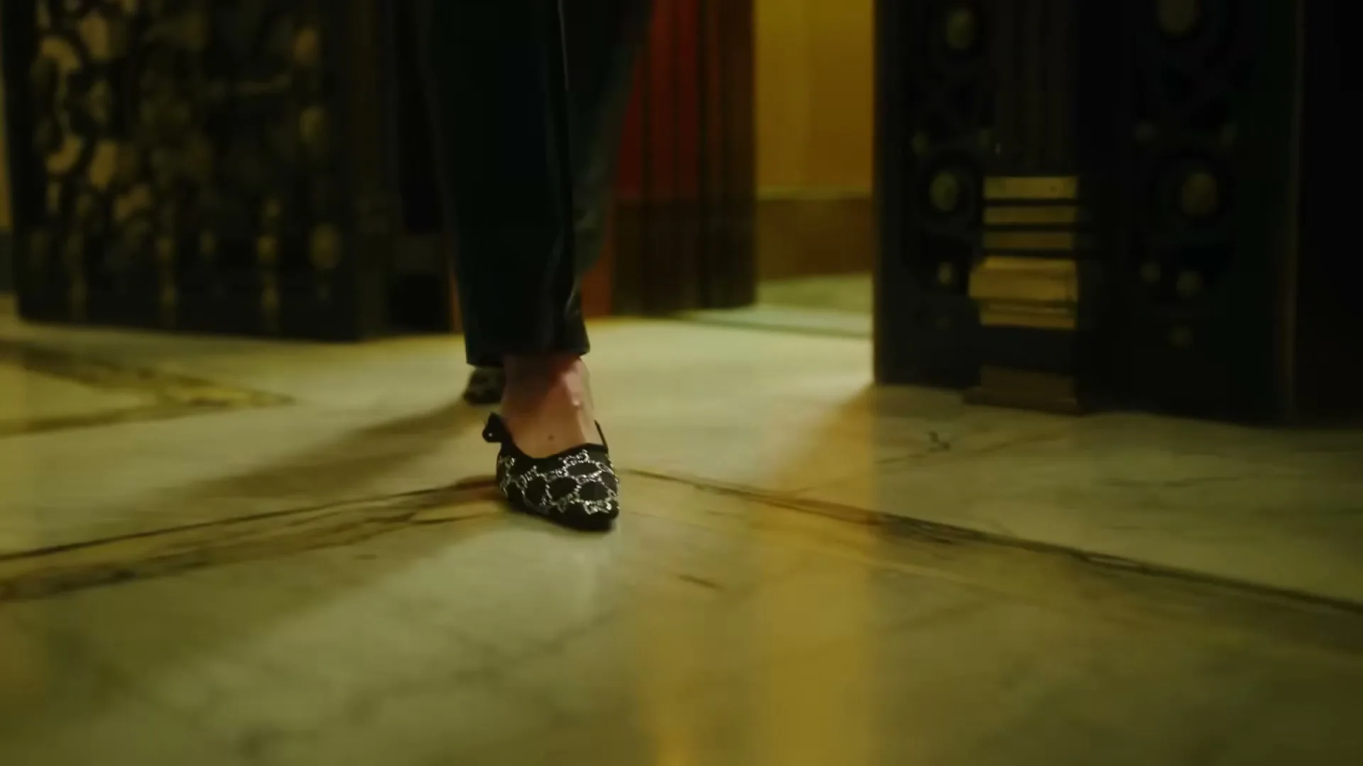 A person wearing black pants and black patterned mules walks across a marble floor, with ornate dark doors on either side.