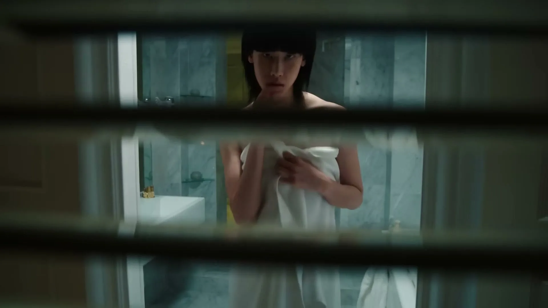 A person with a bob haircut, wrapped in a white towel, looks directly at the camera through venetian blinds in a luxurious bathroom.