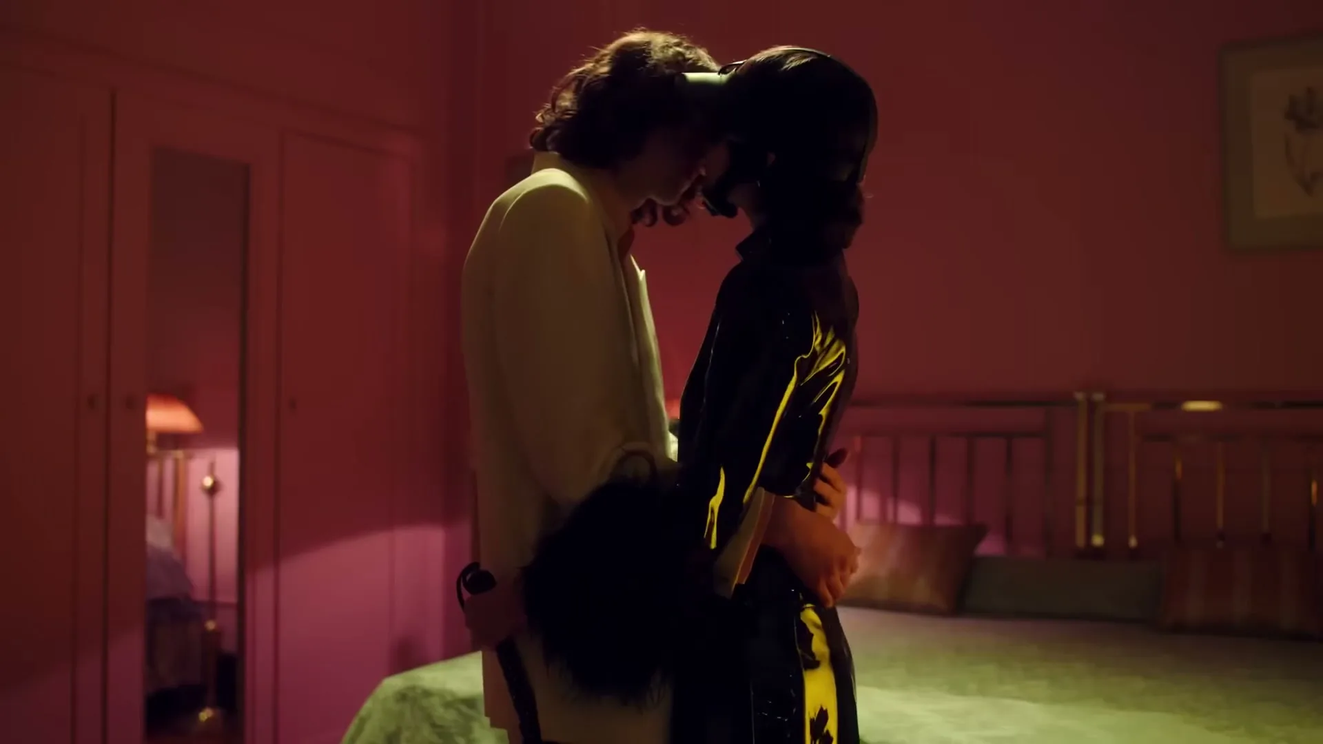 A man in a white jacket and a woman in a black outfit with a headpiece embrace intimately in a dimly lit, pink-walled bedroom.