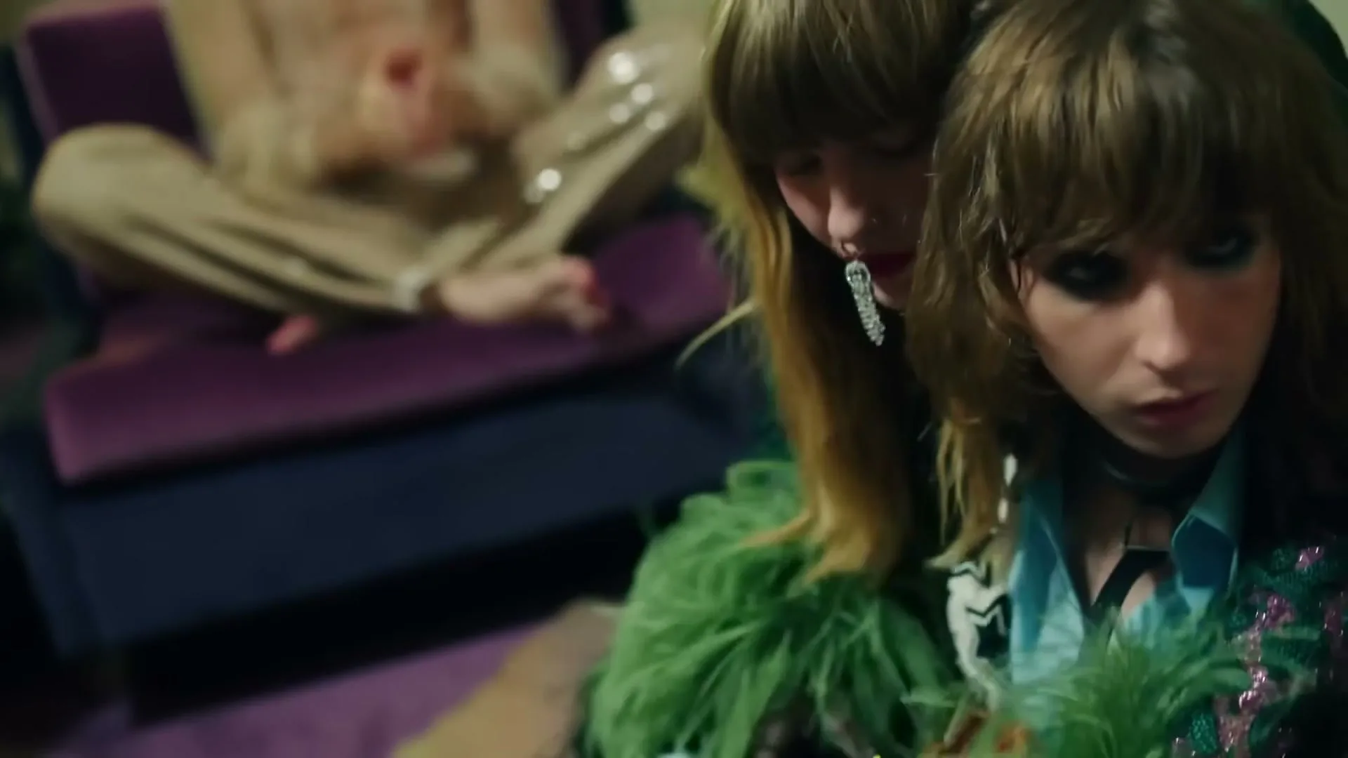 Two young people with long hair and dramatic makeup embrace, one wearing a green feathered boa, in a dimly lit room.