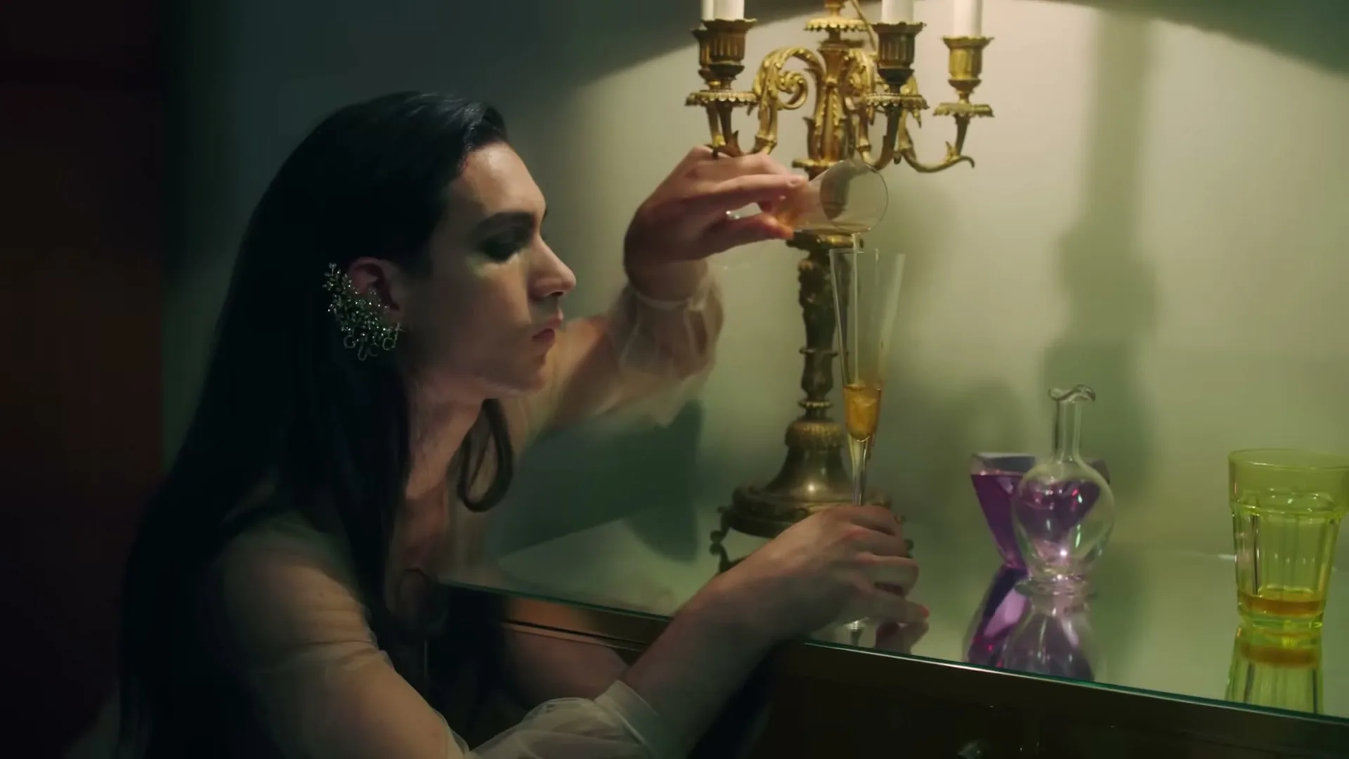 A person with long dark hair and an ornate ear cuff pours liquid from a small glass into a champagne flute next to a candelabra on a mirrored surface.
