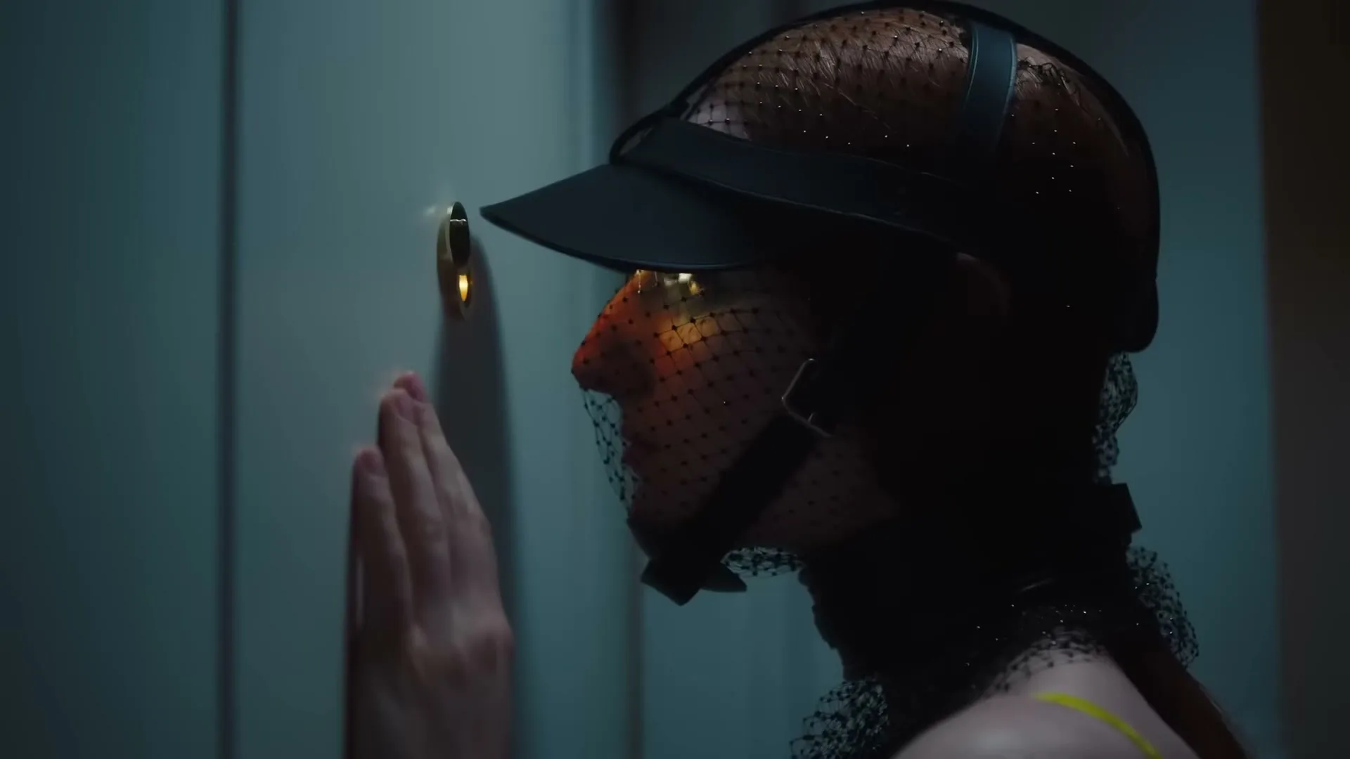 A person with a veiled face and a cap peers through a keyhole, with light emanating from the other side of the door.