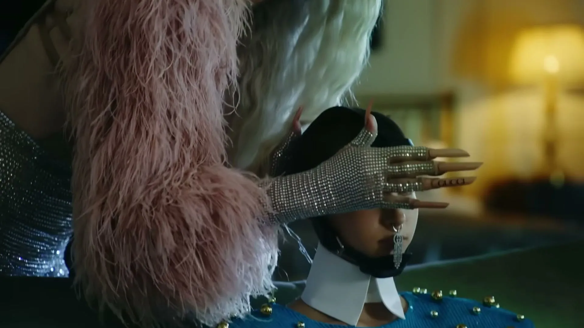 A person with long blonde hair and a pink feathered coat covers the eyes of another person wearing a blue studded top and a nose ring, in a dimly lit room.