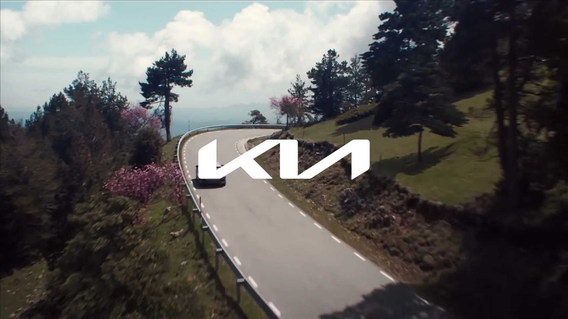 A Kia EV6 drives along a winding mountain road, with the Kia logo superimposed over the scene, under a partly cloudy sky.