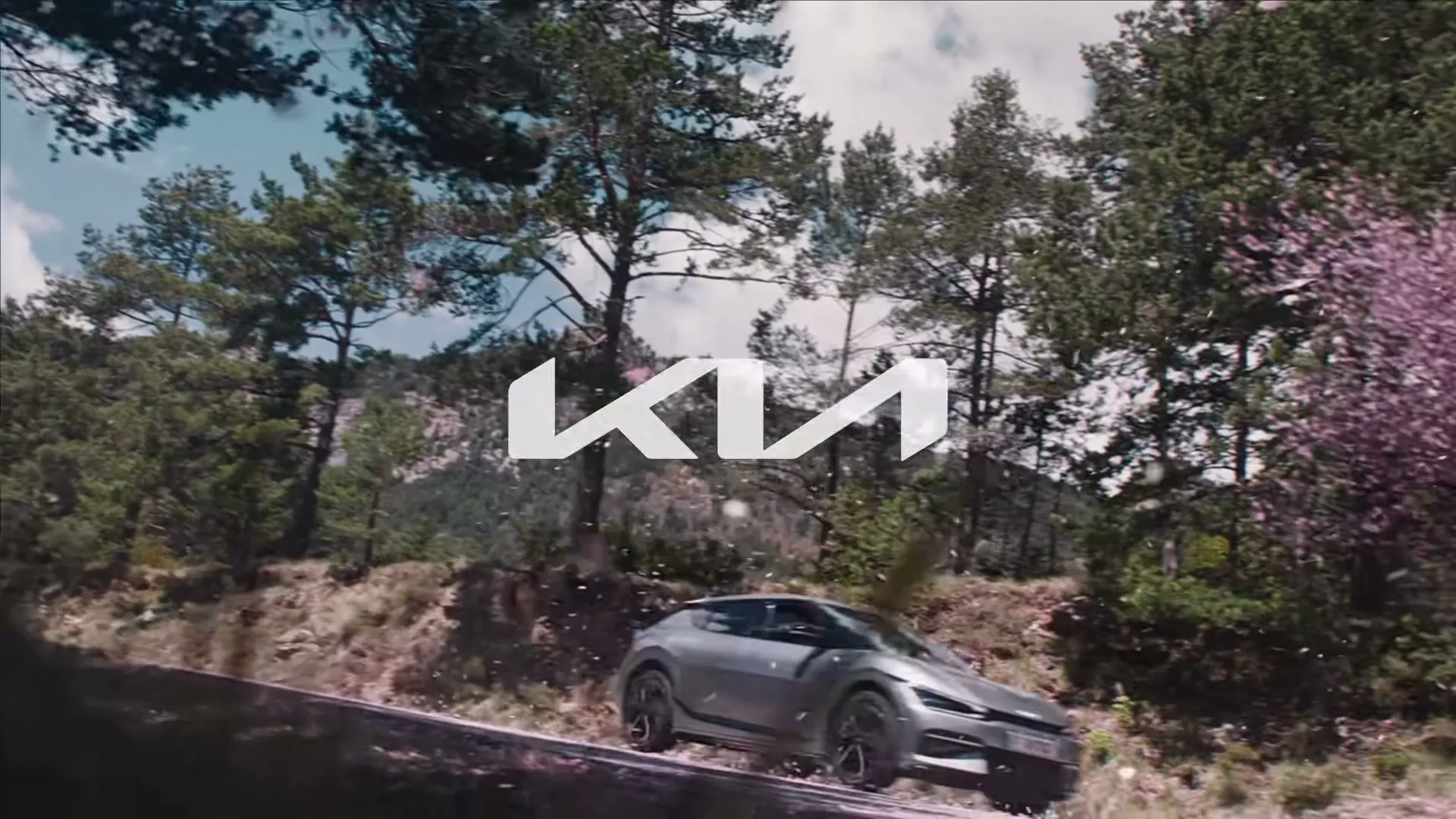 A silver Kia EV6 drives on a winding mountain road, surrounded by pine trees and falling pink petals, with the Kia logo superimposed.