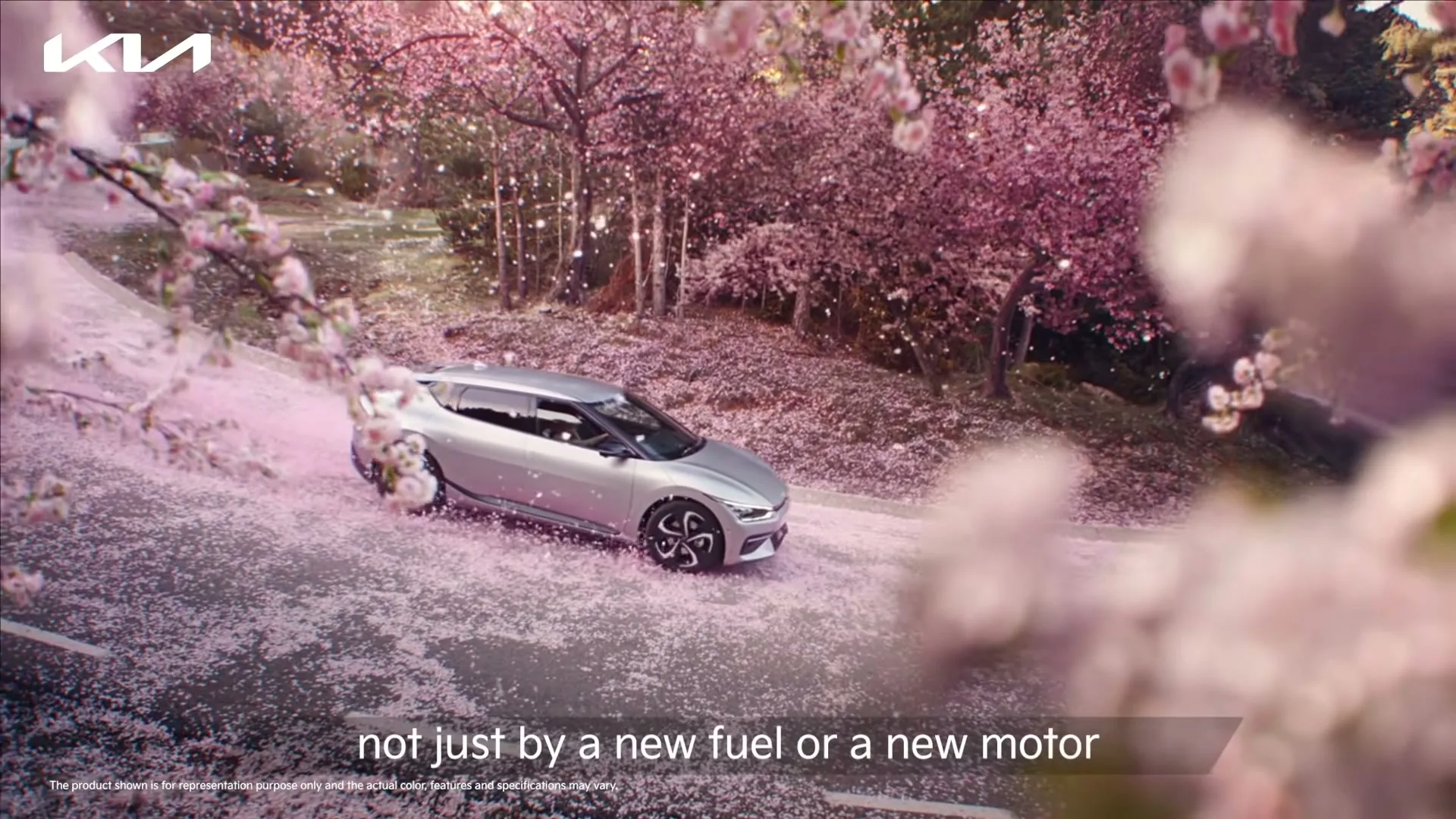 A silver Kia EV6 drives on a winding road covered in pink cherry blossom petals, surrounded by blooming cherry trees on a sunny day.