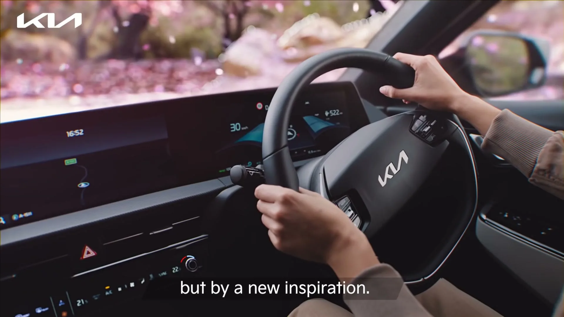A person's hands are on the steering wheel of a Kia EV6, driving through a scenic route with pink petals falling.