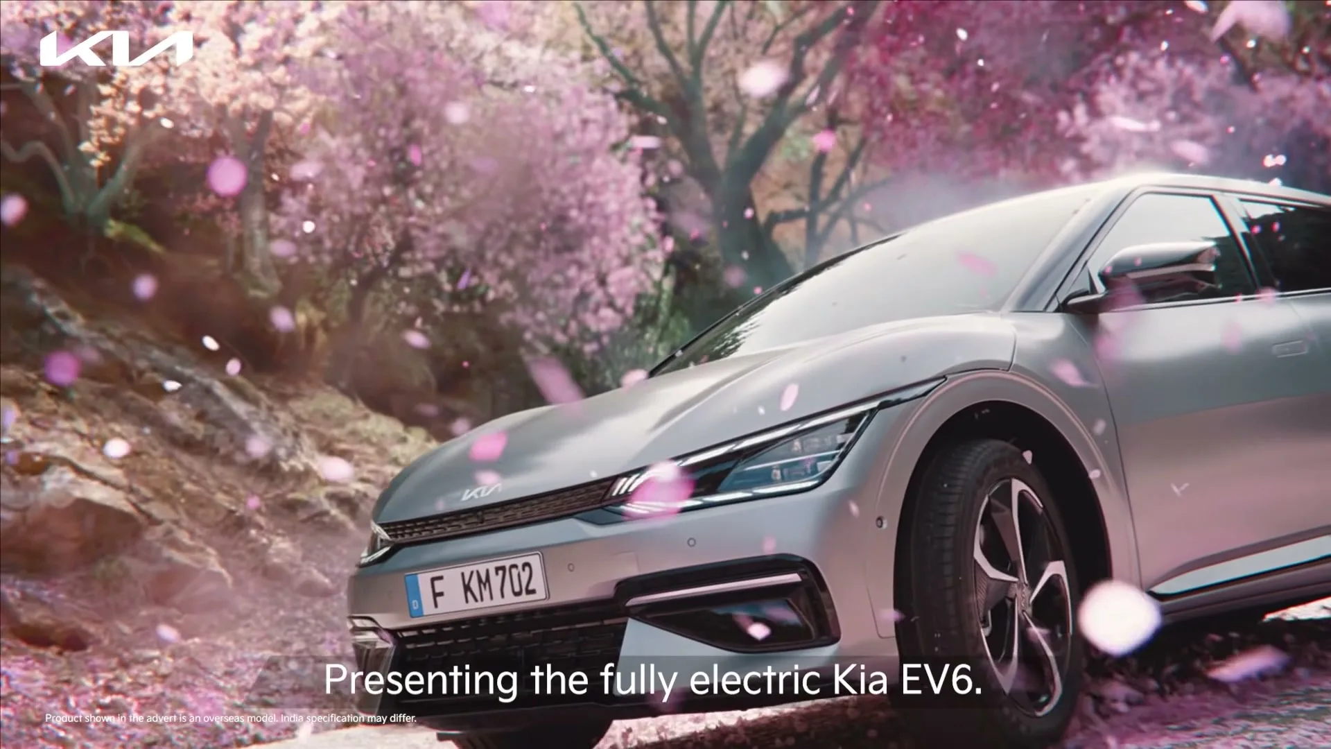 A silver Kia EV6 is parked on a dirt road, surrounded by falling pink cherry blossoms, with a forest of cherry trees in the background.