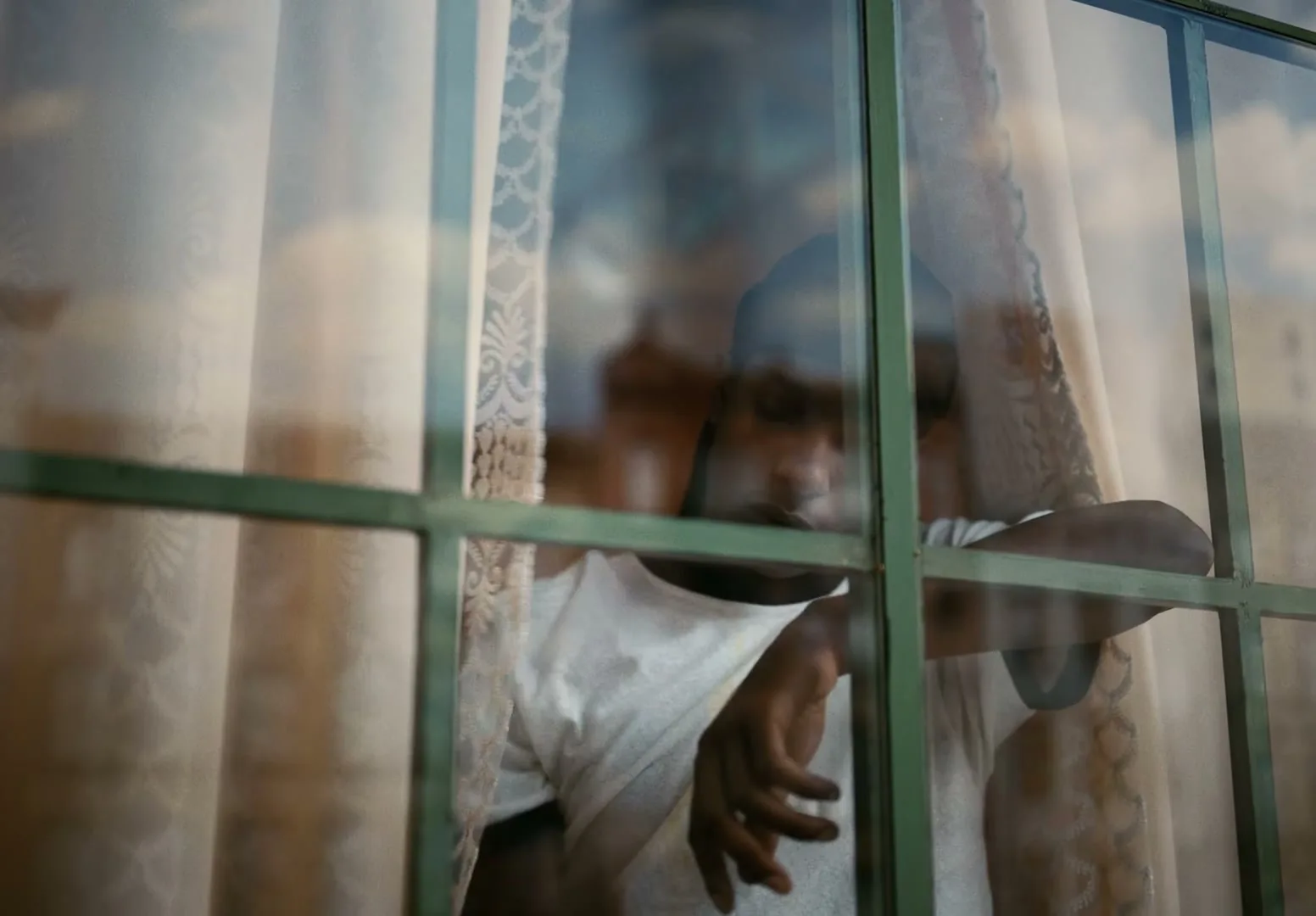 A young man with a durag and white t-shirt looks out a window with green panes, his reflection visible, as the camera slowly zooms out.