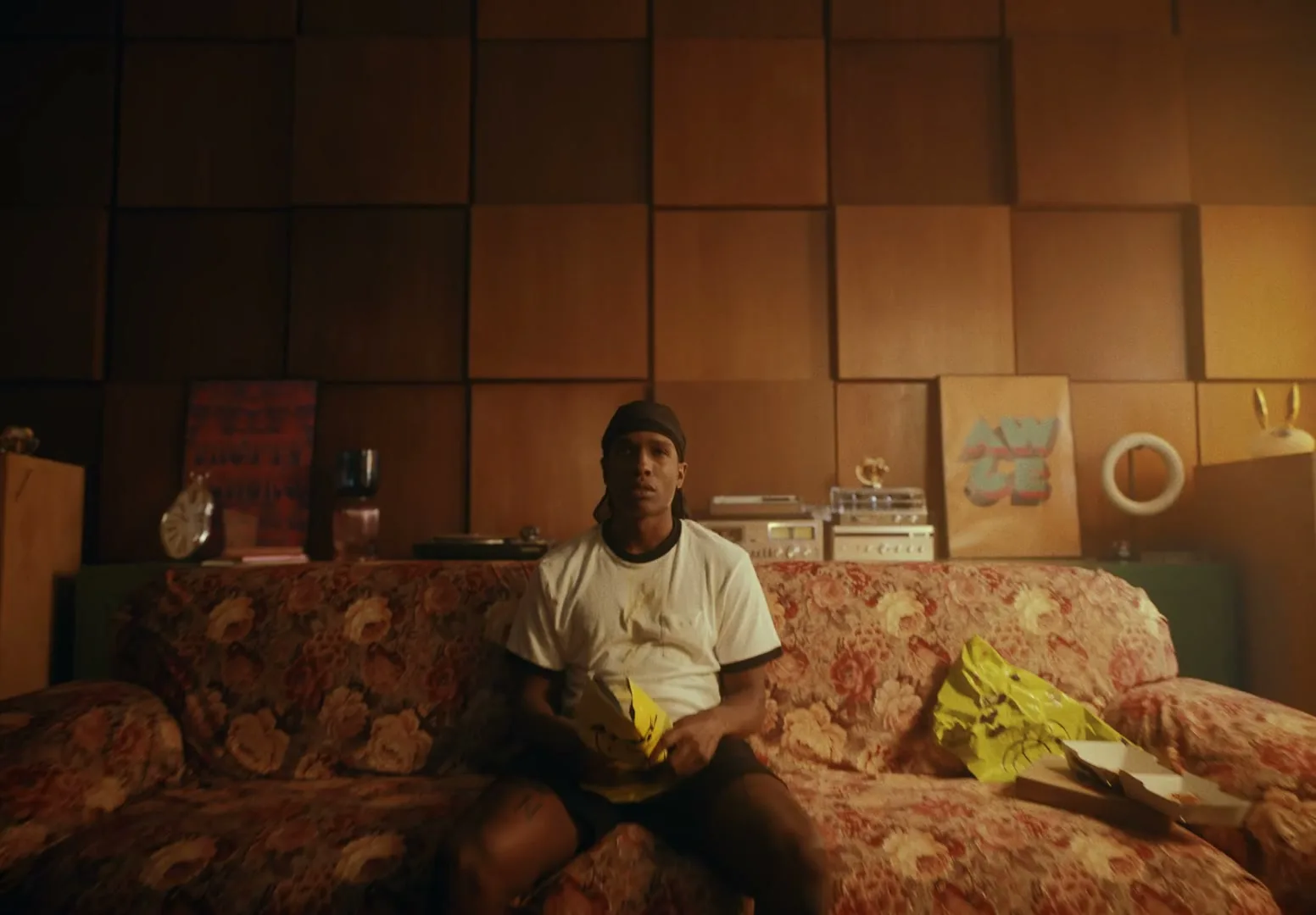A young man in a durag and t-shirt sits on a floral couch in a messy living room, looking up as the camera pulls back.