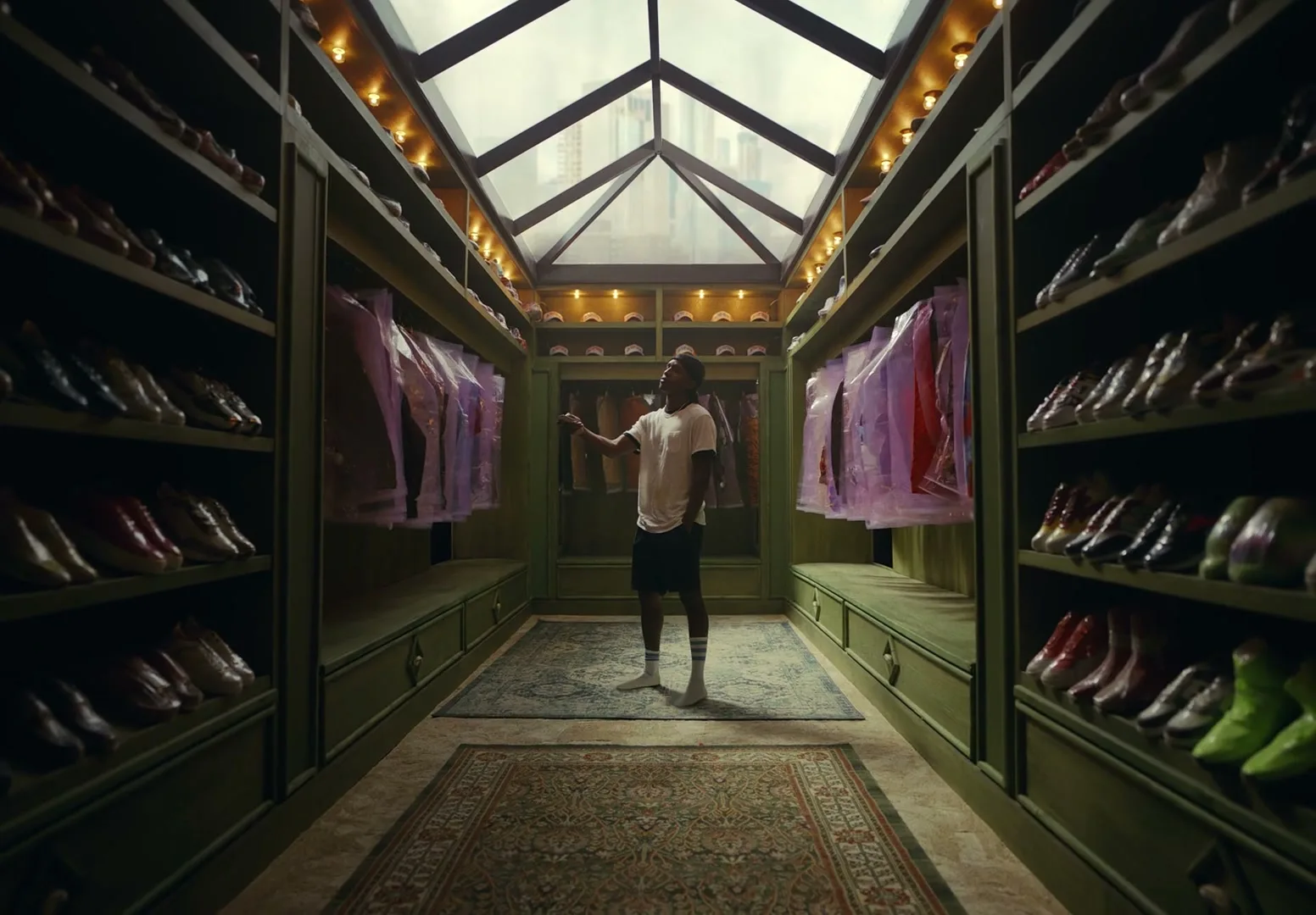 A man in a white t-shirt and shorts stands in a luxurious walk-in closet, surrounded by shoes and clothes, looking at a garment.