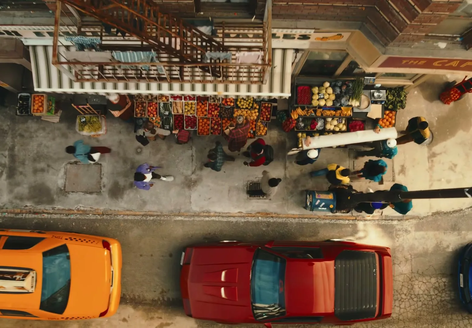 An overhead shot of a busy street market with people walking, cars parked, and a fruit stand, as two men carry a large roll.
