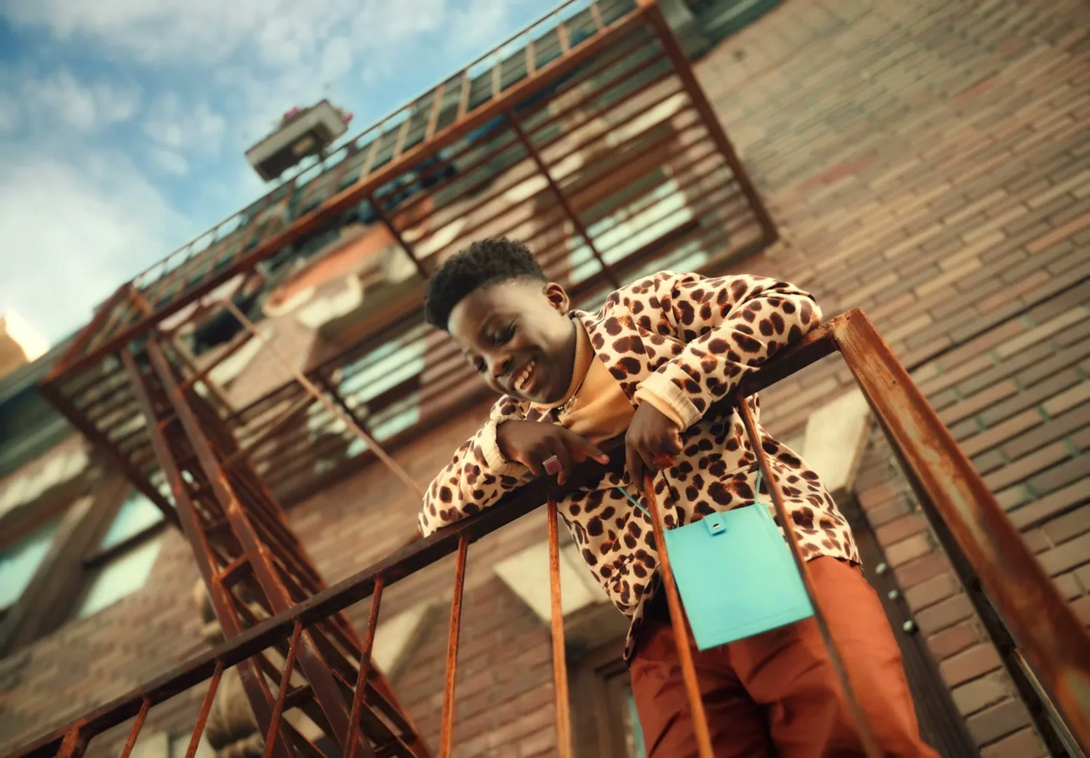 A young person in a leopard print jacket leans on a rusty fire escape railing, looking down and smiling at the camera on a sunny day.
