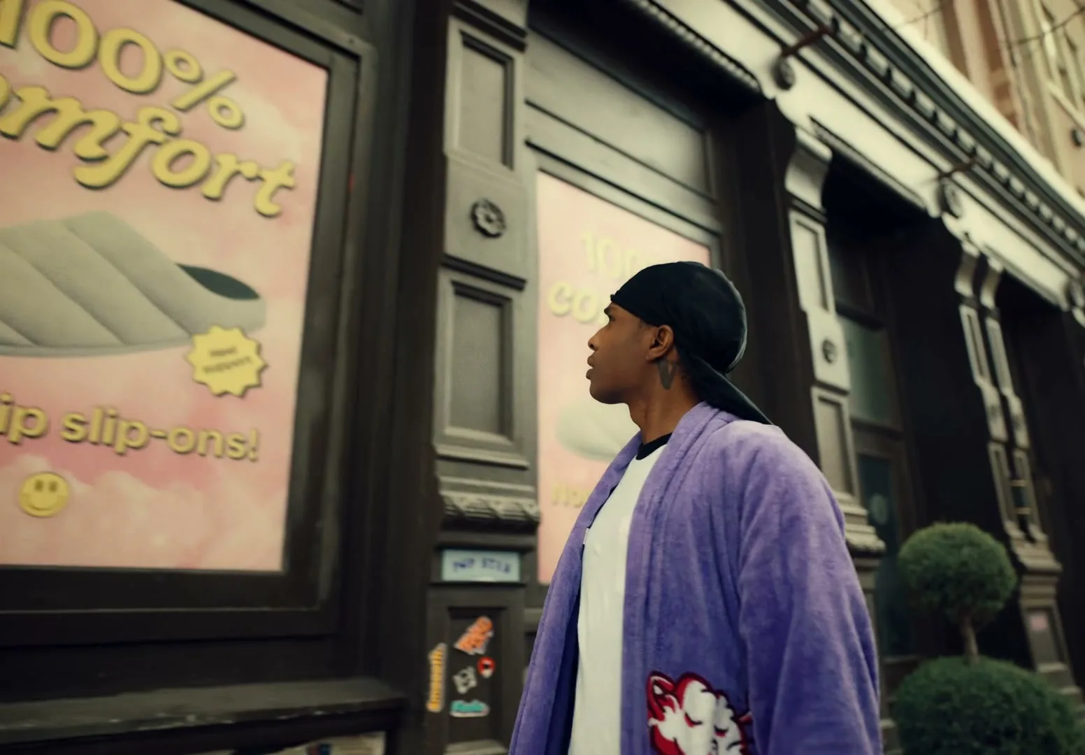 A$AP Rocky, wearing a purple robe and black durag, looks at a store window displaying a slipper advertisement on a city street.