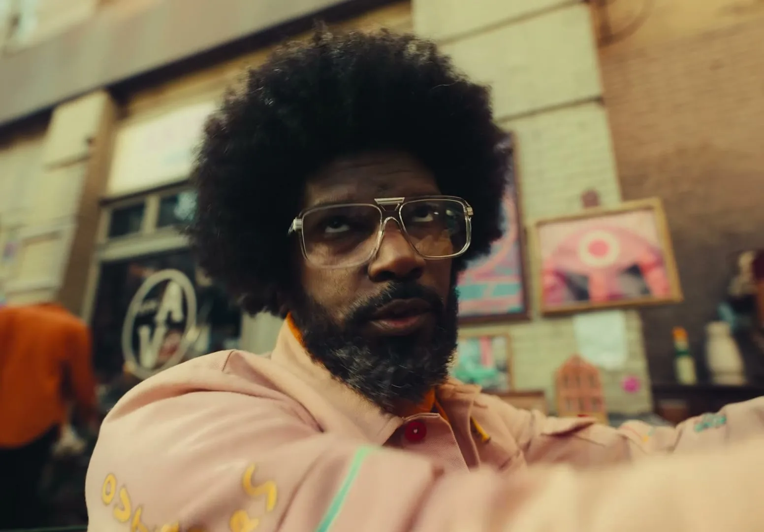 A man with an afro and beard, wearing glasses and a pink jacket, looks around with a surprised expression in an outdoor market.