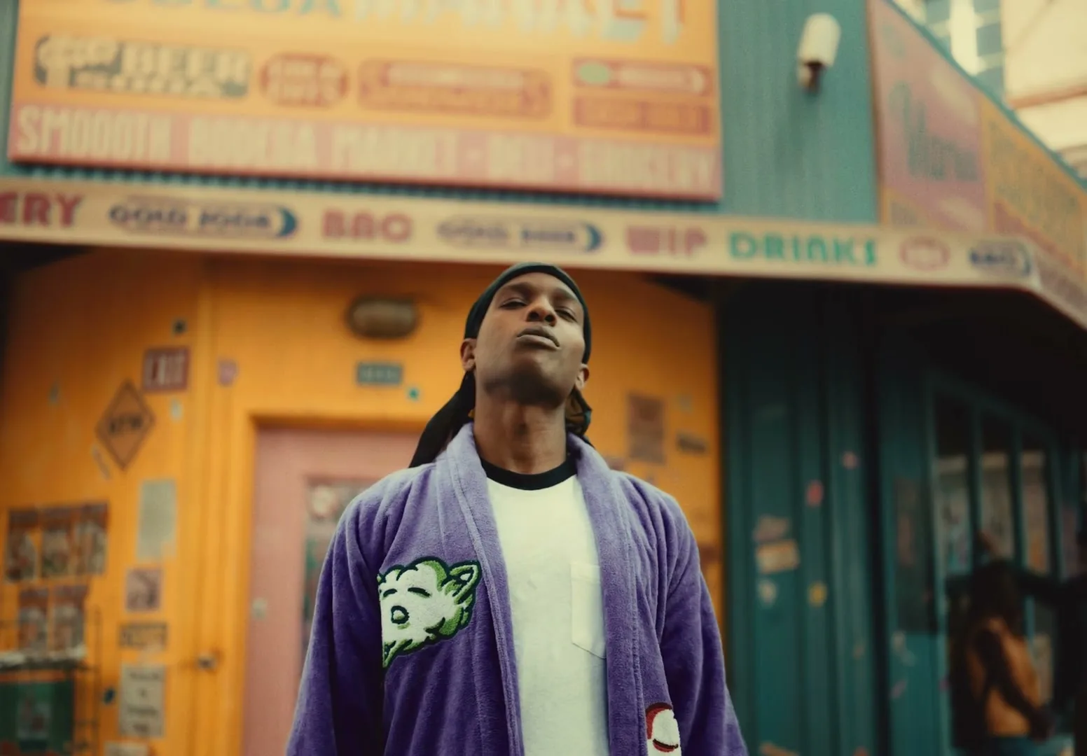 A$AP Rocky, a Black man in his 30s, wearing a purple robe and black durag, looks up and then forward, standing in front of a colorful bodega.