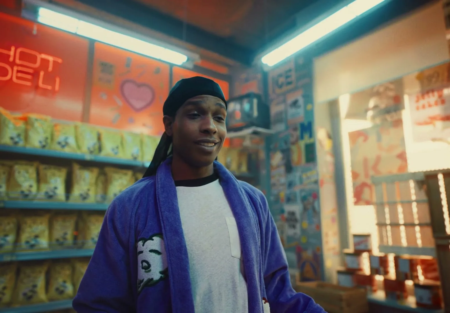 A$AP Rocky, a Black man in his 30s, wearing a purple robe and black durag, stands in a brightly lit convenience store, looking around with a neutral expression.