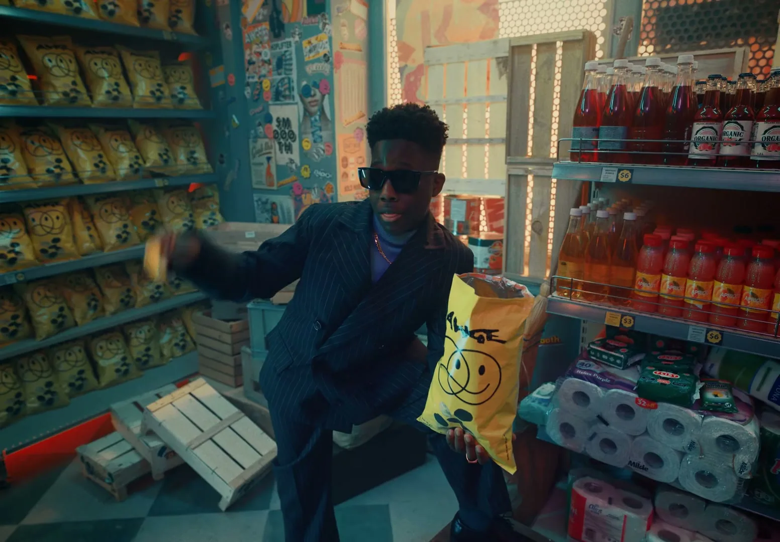 A$AP Rocky, a Black man in his 30s, wearing sunglasses and a black coat, smiles with gold grills in a grocery store aisle.
