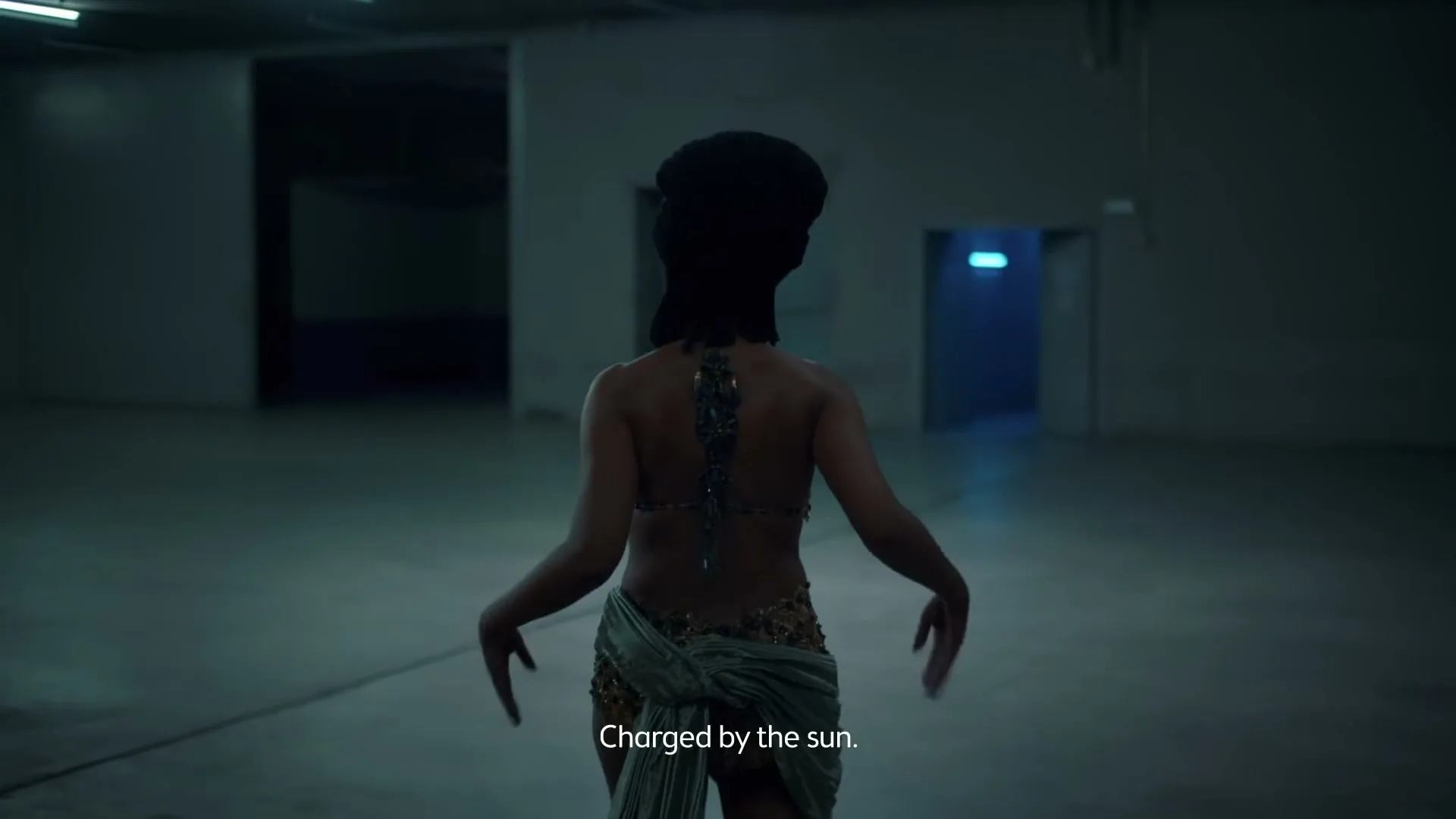 A person with dark skin and a black head covering walks away from the camera in a dimly lit, empty warehouse, with glowing blue light visible through a distant doorway.