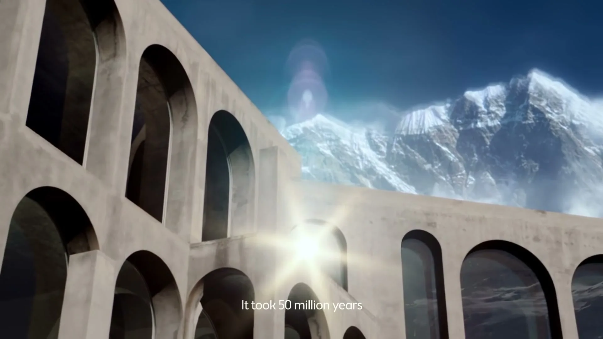 A concrete building with arched windows stands in the foreground, with snow-capped mountains under a blue sky in the background, as sunlight flares through an archway.