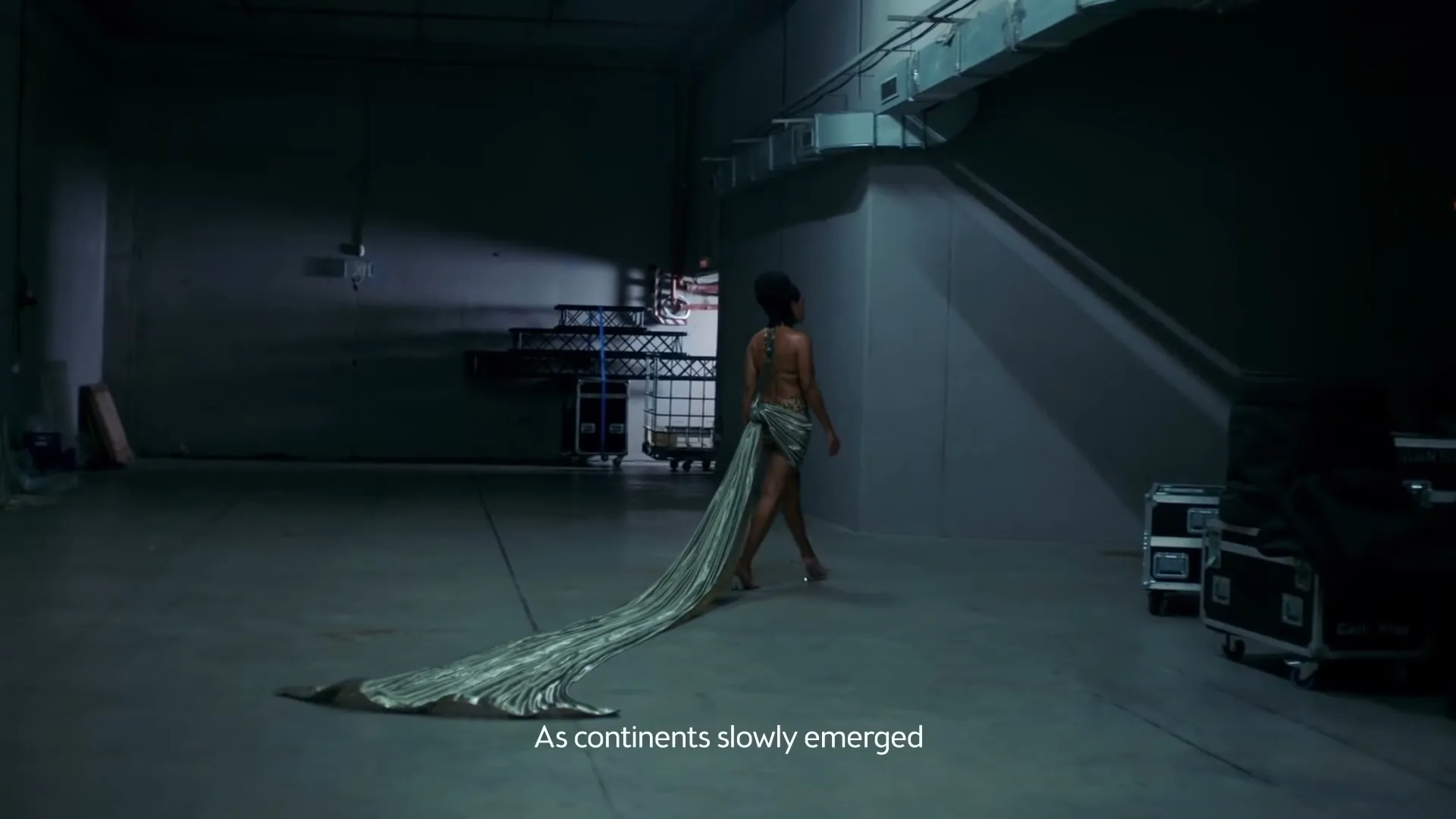 A woman in a long, flowing silver dress walks away from the camera in a dimly lit, empty warehouse, with industrial equipment visible in the background.