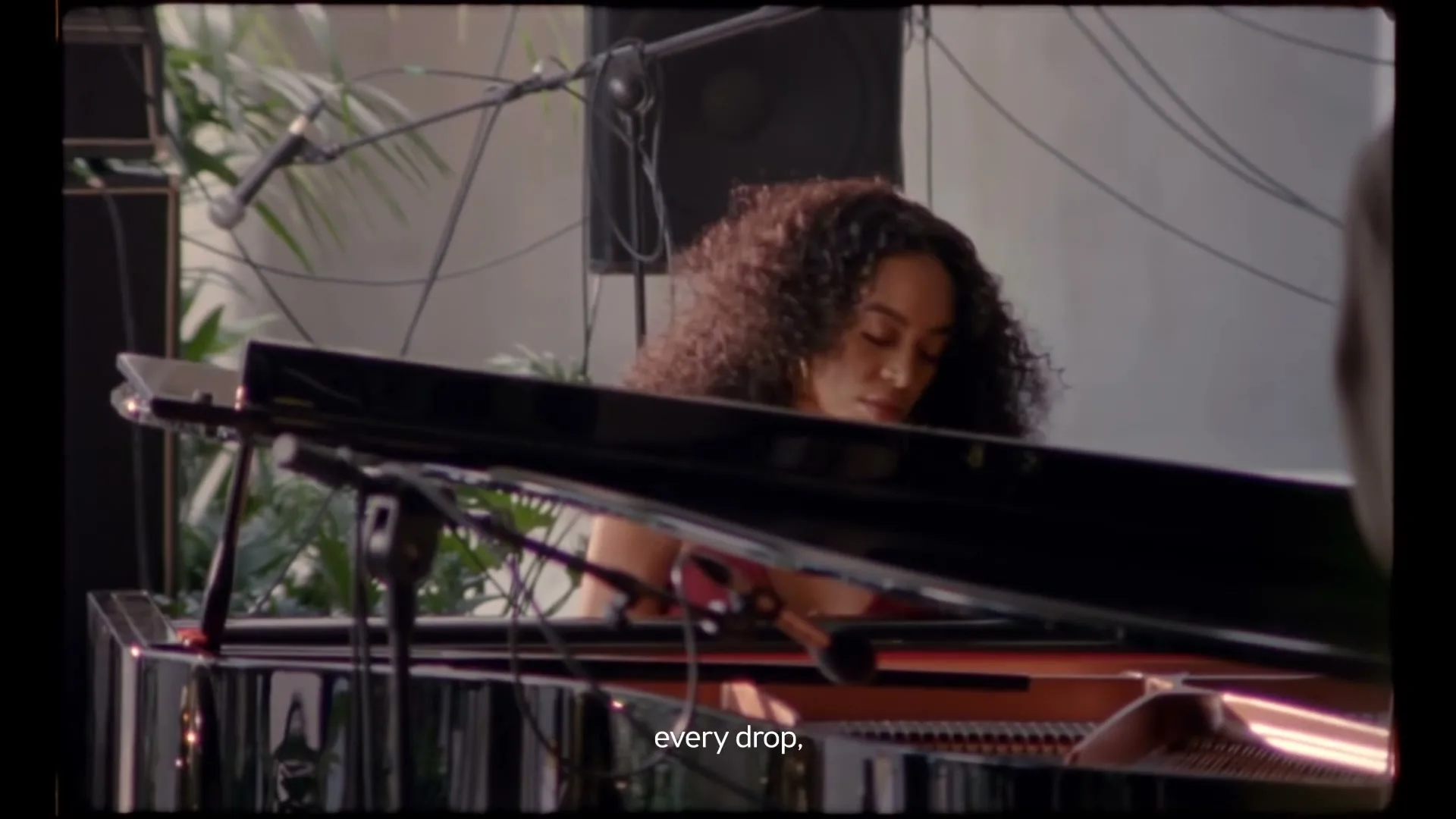 A woman with curly hair plays a grand piano, looking focused in a bright, airy room with plants and music equipment.