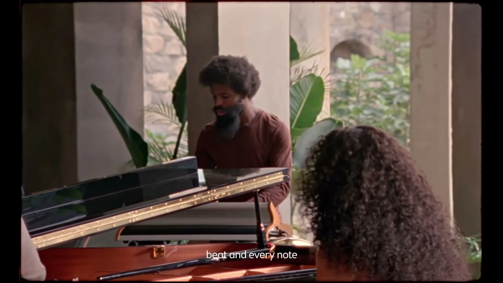 A man with a beard and afro plays a grand piano while a woman with curly hair sits beside him, in a luxurious, open-air setting.