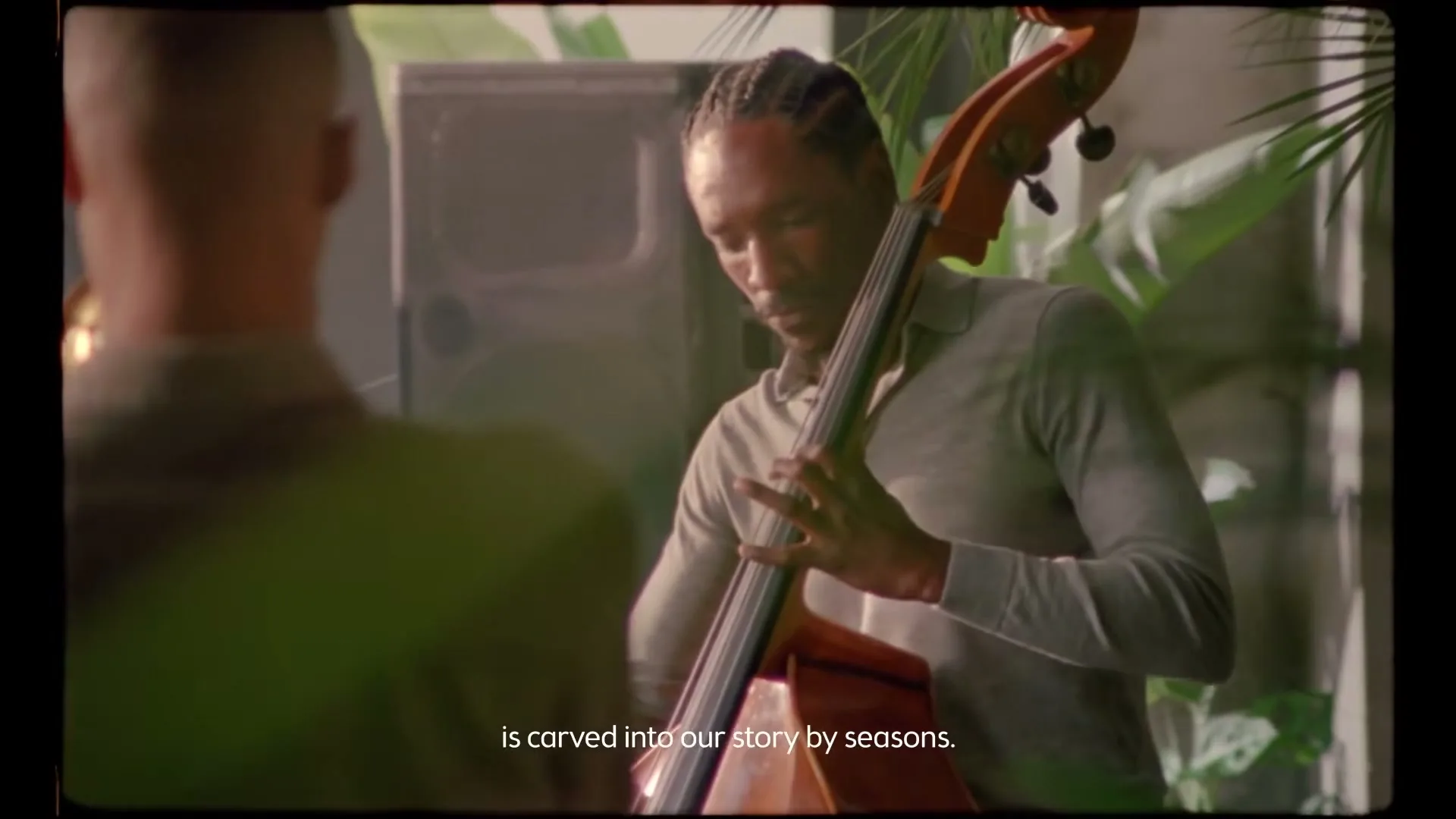 A Black man with dreadlocks plays an upright bass, seen over the shoulder of another man, in a lush, dimly lit interior setting.