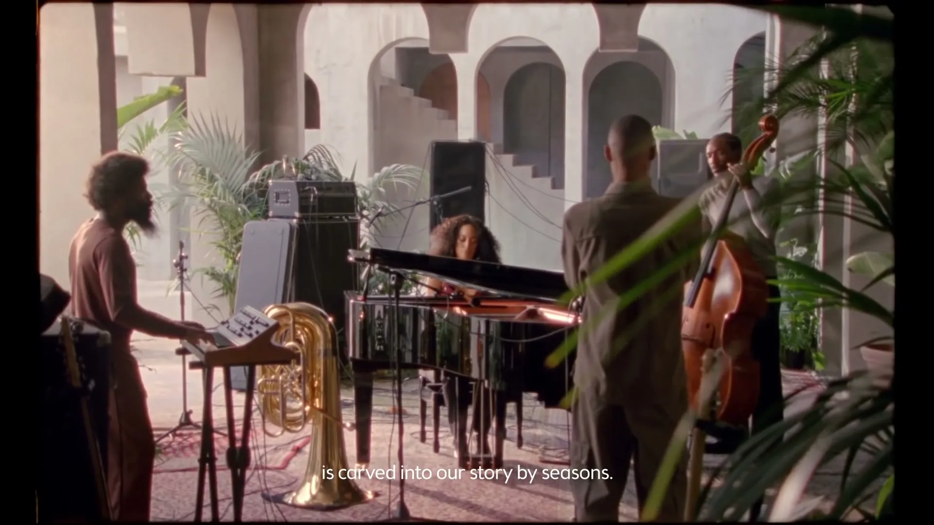 A diverse jazz band performs in a beautiful, sunlit courtyard with arched doorways and lush greenery, featuring a female pianist and male musicians.