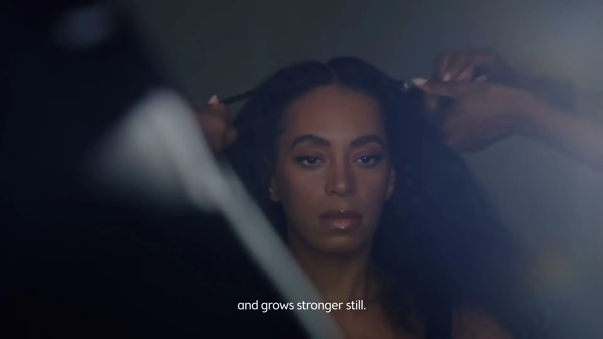 A woman with long dark hair sits as two people style her hair, with a soft light on her face in a dark room.