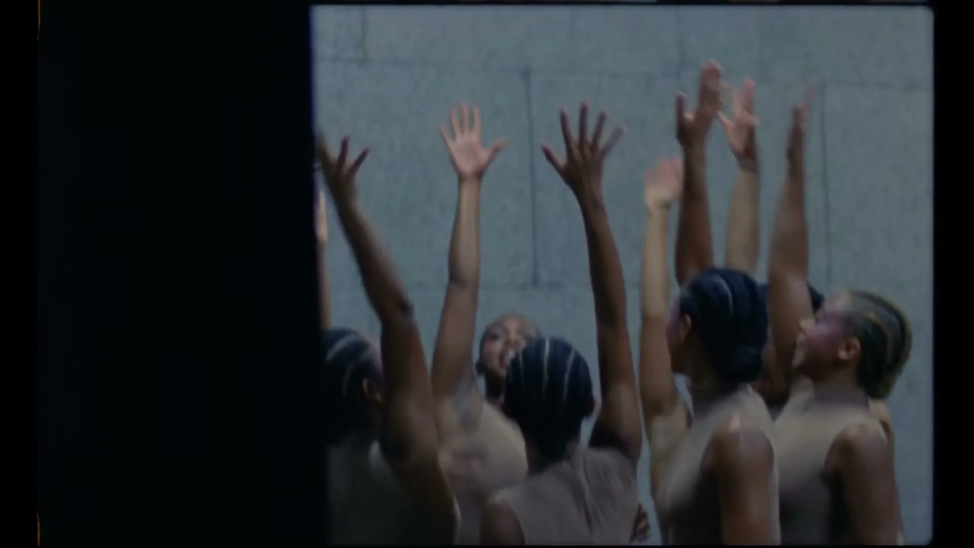 A group of diverse women in neutral bodysuits huddle together, then raise their hands in unison, in a dimly lit room.