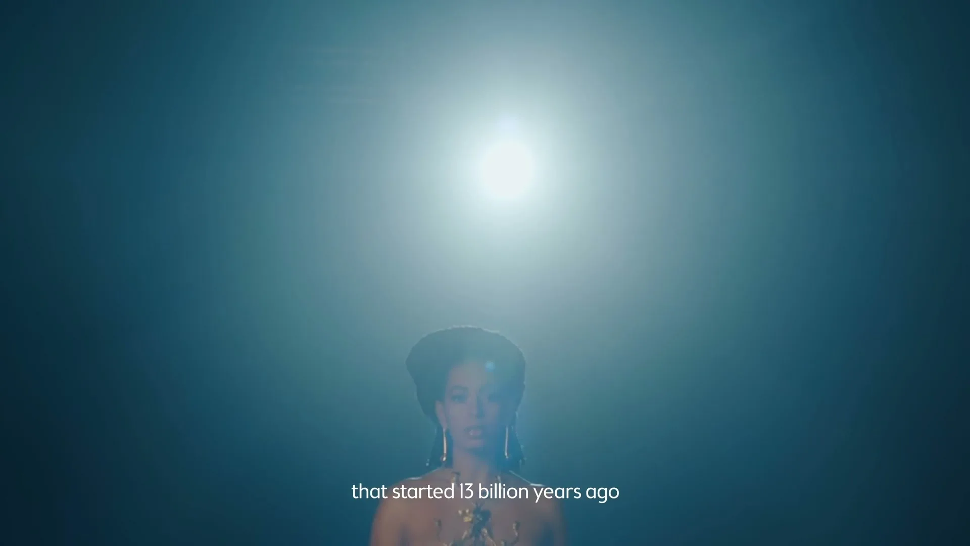 A woman with dark skin and hair, wearing a gold top, looks up at a bright light in a dark, ethereal space.