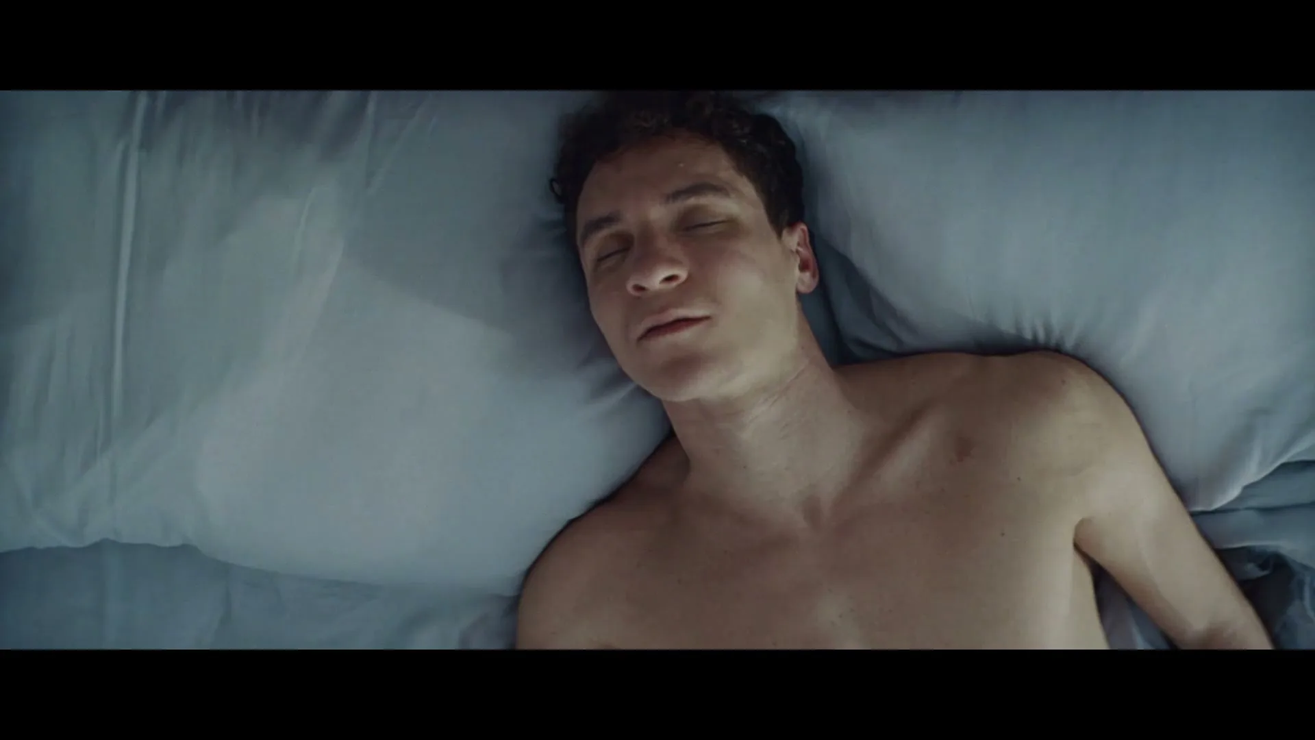 A young man with curly hair lies shirtless in bed, slowly opening his eyes and looking around, in a dimly lit bedroom.