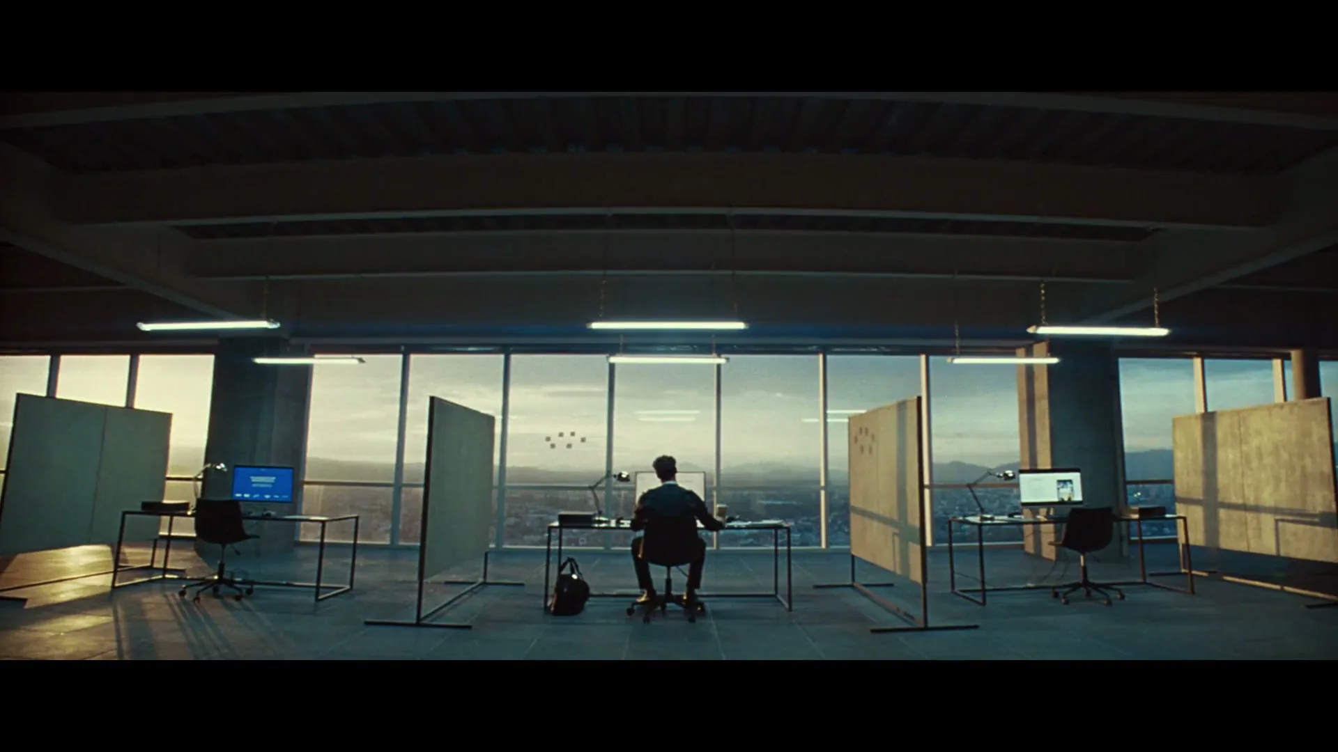 A man sits at a desk in a large, empty office with floor-to-ceiling windows overlooking a city at dusk.
